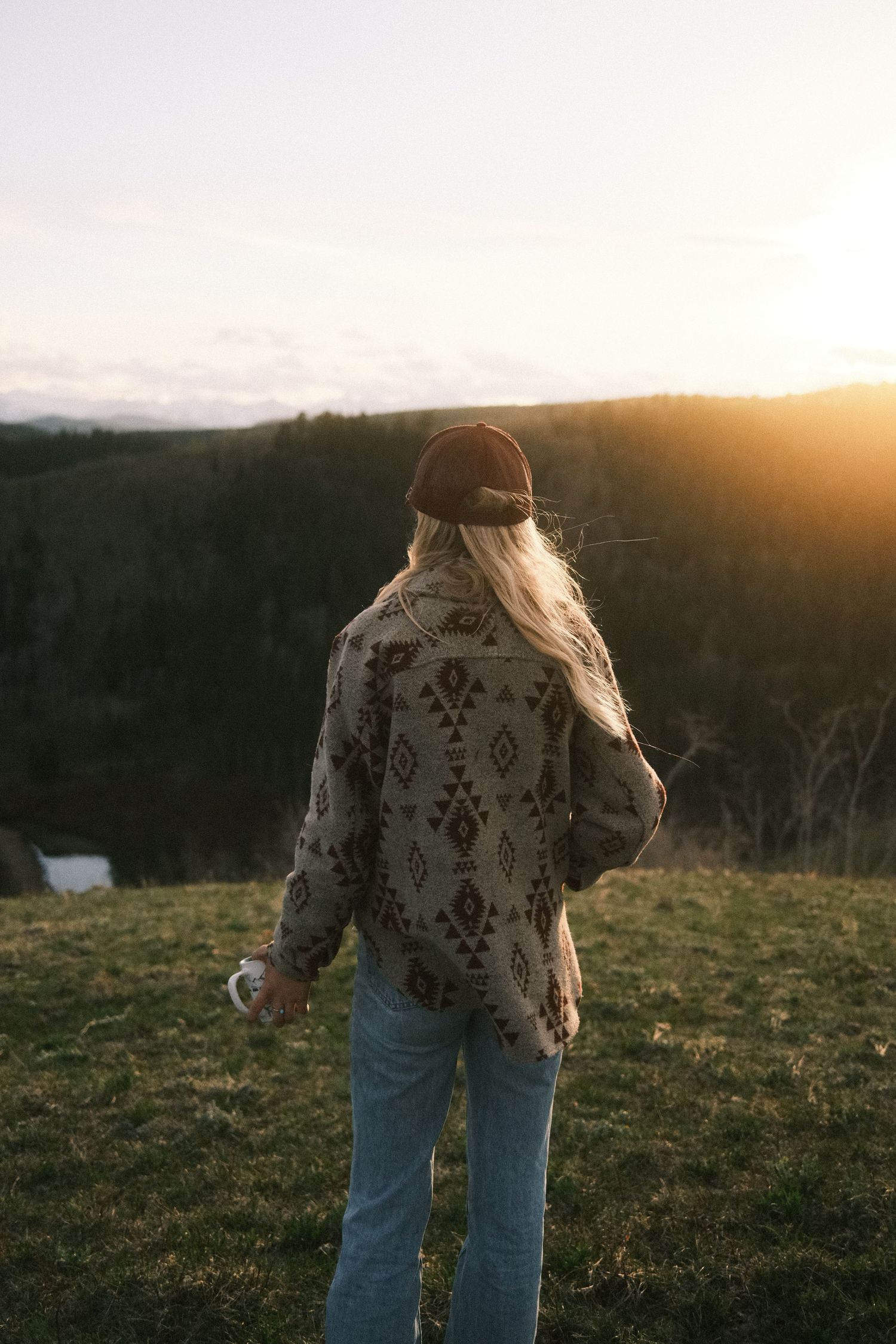 Woman in Aztec print jacket and cap gazes at golden sunset over forested valley landscape.