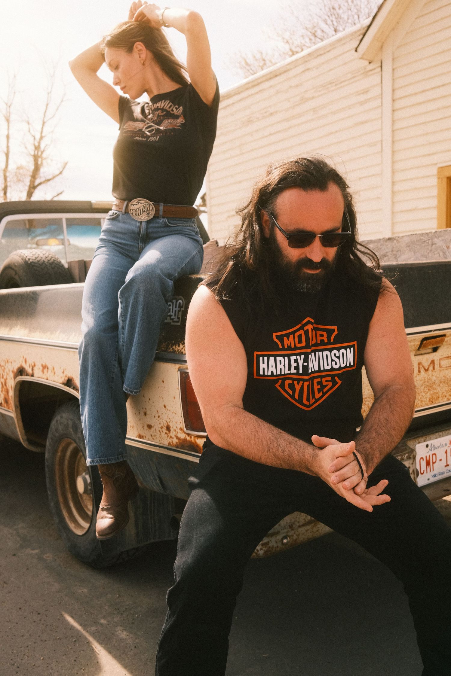 Two people in Harley-Davidson shirts pose on a rusty vintage pickup truck outdoors in casual Western style.