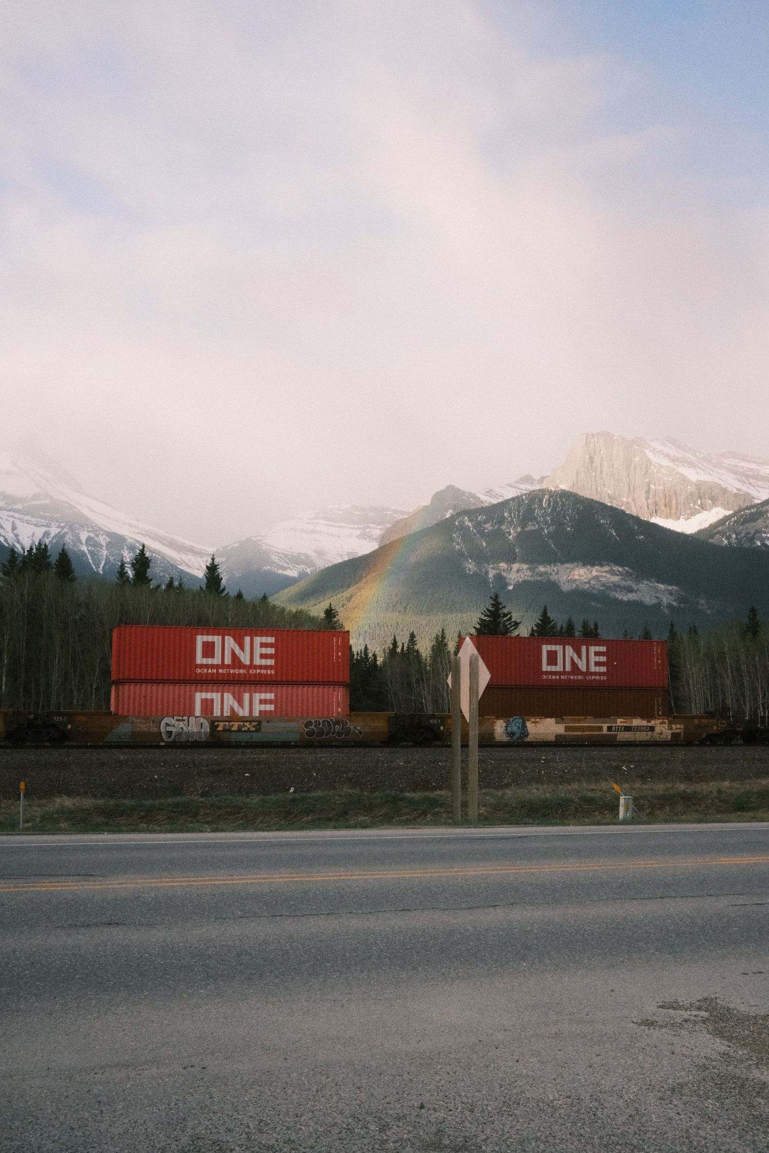 Red ONE shipping containers on a train with a rainbow and snow-capped Rocky Mountains in the background.
