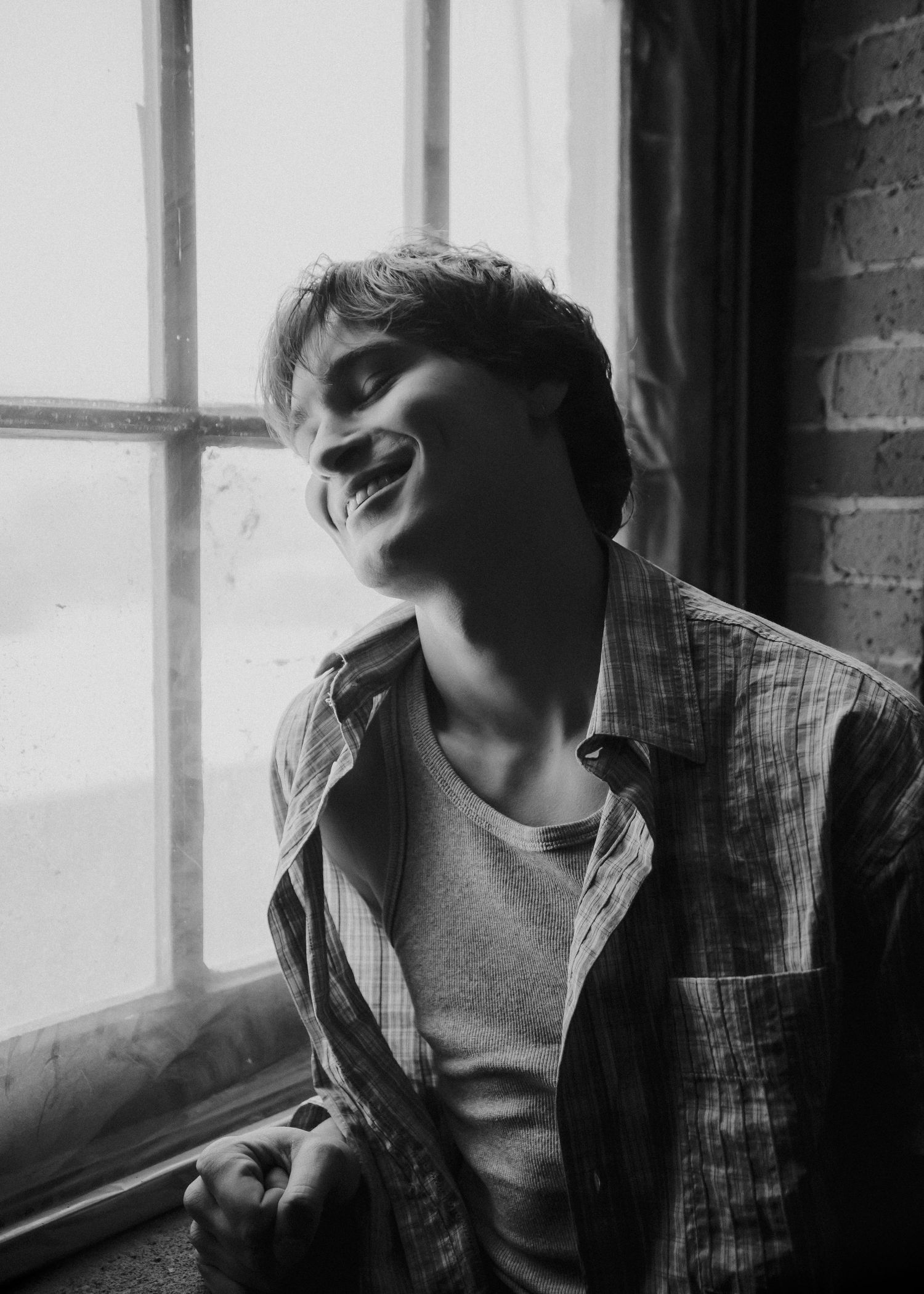 Moody portrait series showing a person in casual attire against a window with brick wall backdrop.