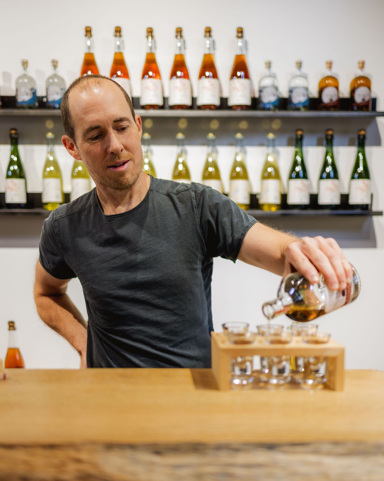 Bartender in black shirt pouring drinks behind wooden counter with liquor bottles displayed.