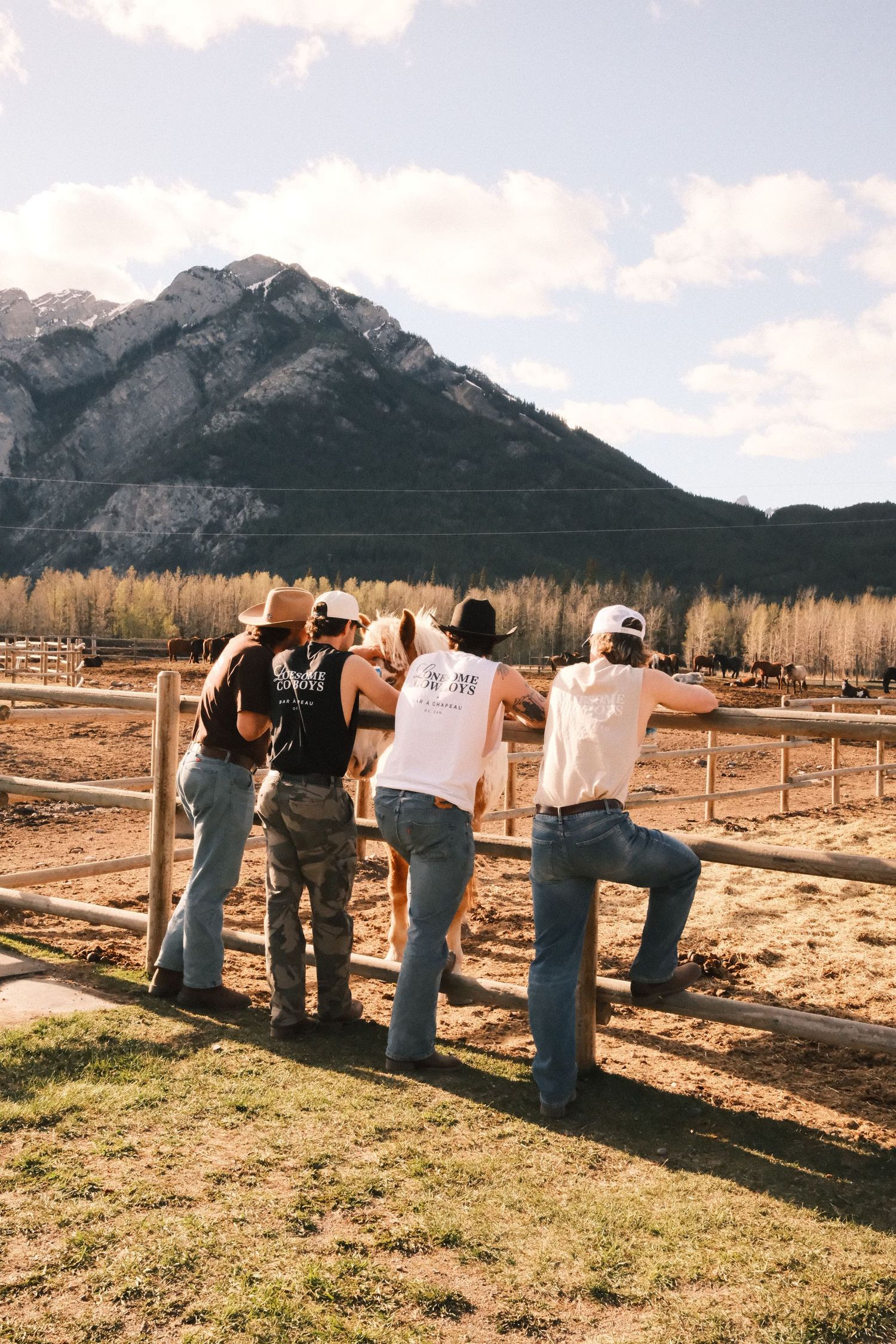 Four people lean on a wooden fence watching a horse with mountains and autumn trees in the background.