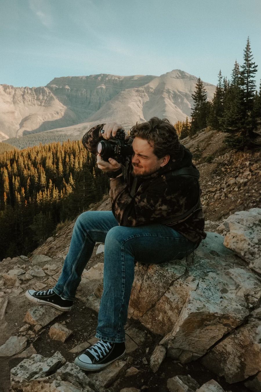 A photographer sits on rocky terrain capturing mountain views with a camera, surrounded by pine trees and peaks.