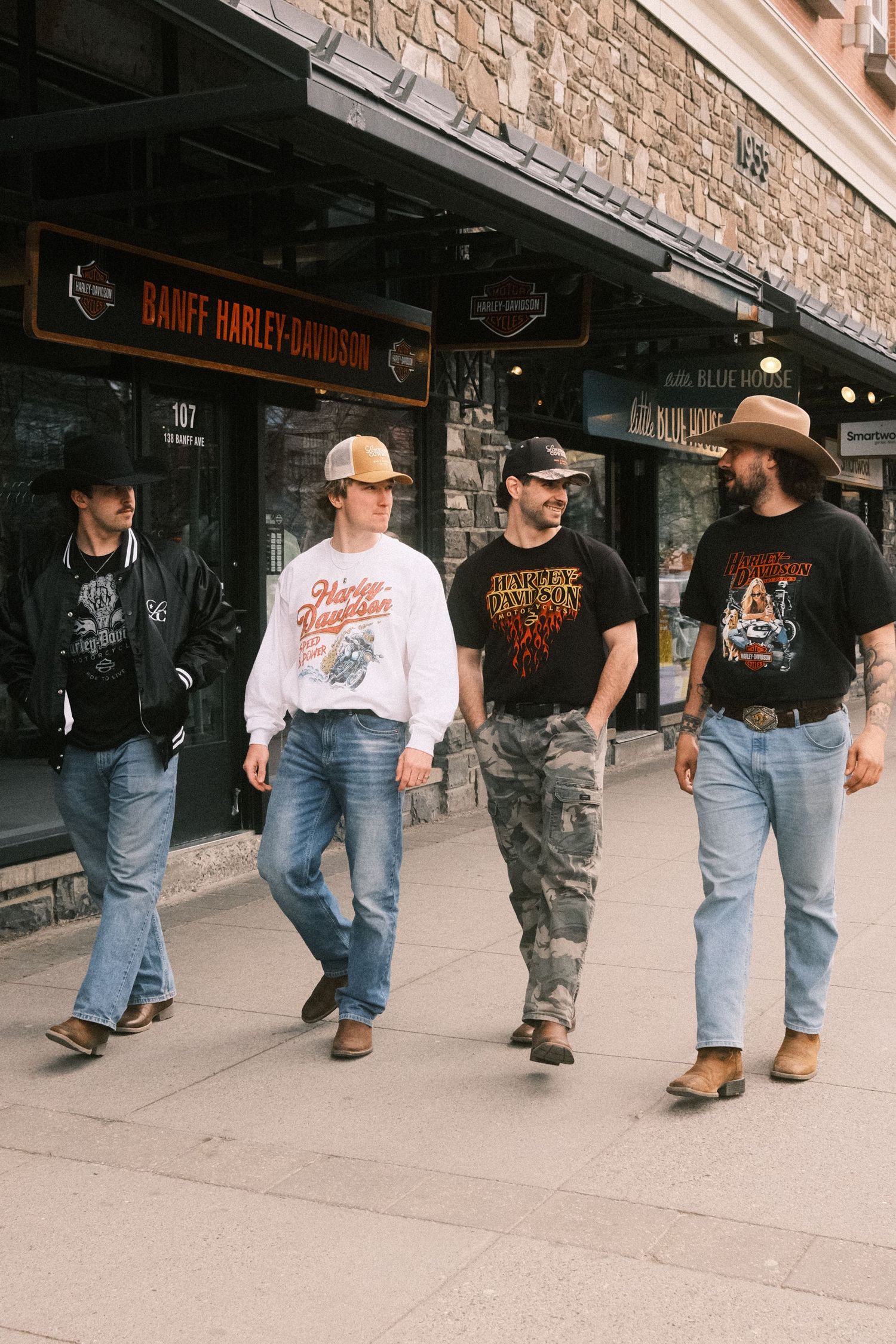 Four men in cowboy hats and Harley-Davidson tees walk past Banff Harley-Davidson store on a sidewalk.