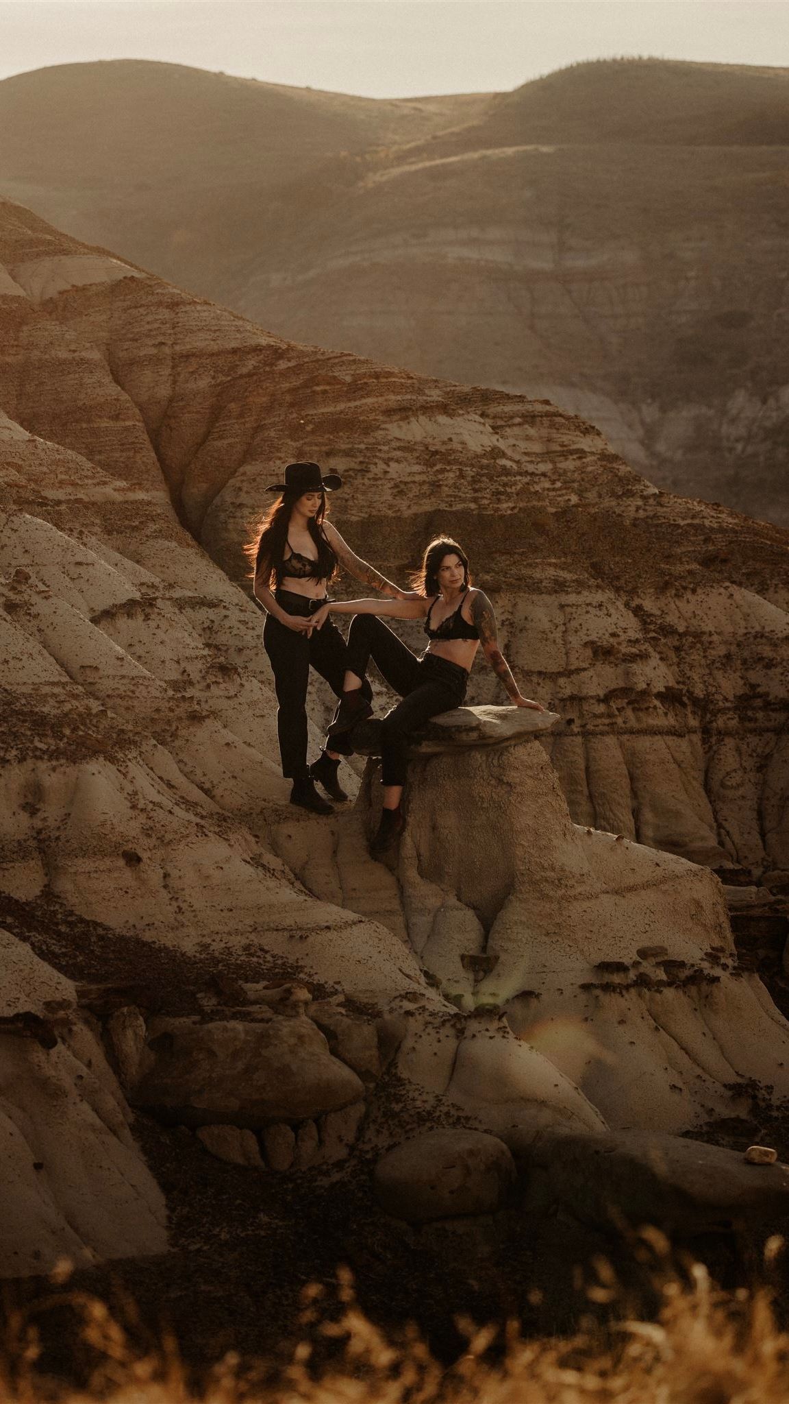 Two women pose on a rocky desert canyon formation at golden hour with layered sandstone hills in the background.