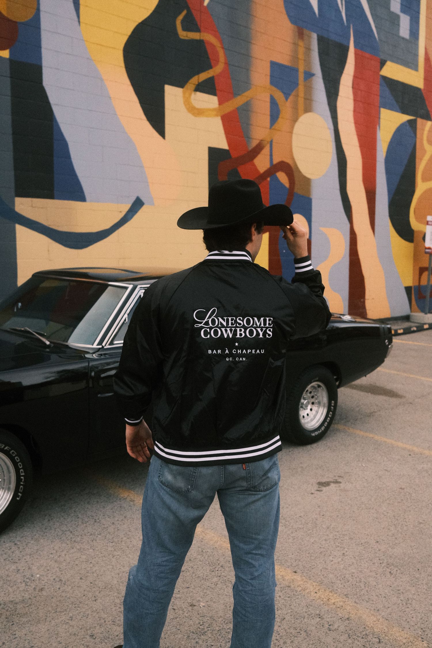 Man in black cowboy hat and Lonesome Cowboys jacket stands by a classic black car in front of a colorful mural.