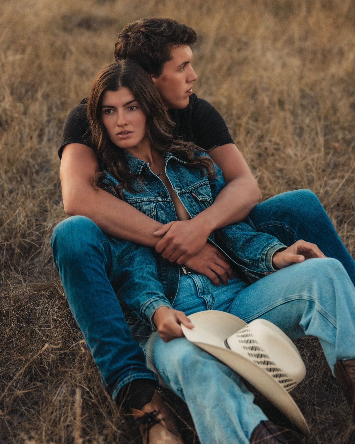 A couple in denim sits together in a field during golden hour, holding a white cowboy hat.