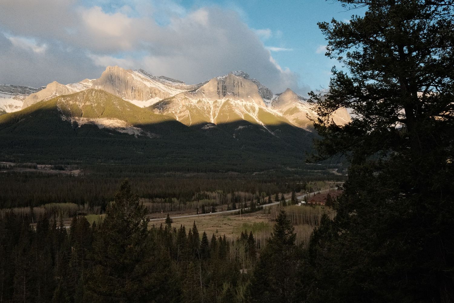 Snow-capped Rocky Mountain peaks rise above a forested valley under dramatic clouds at golden hour.