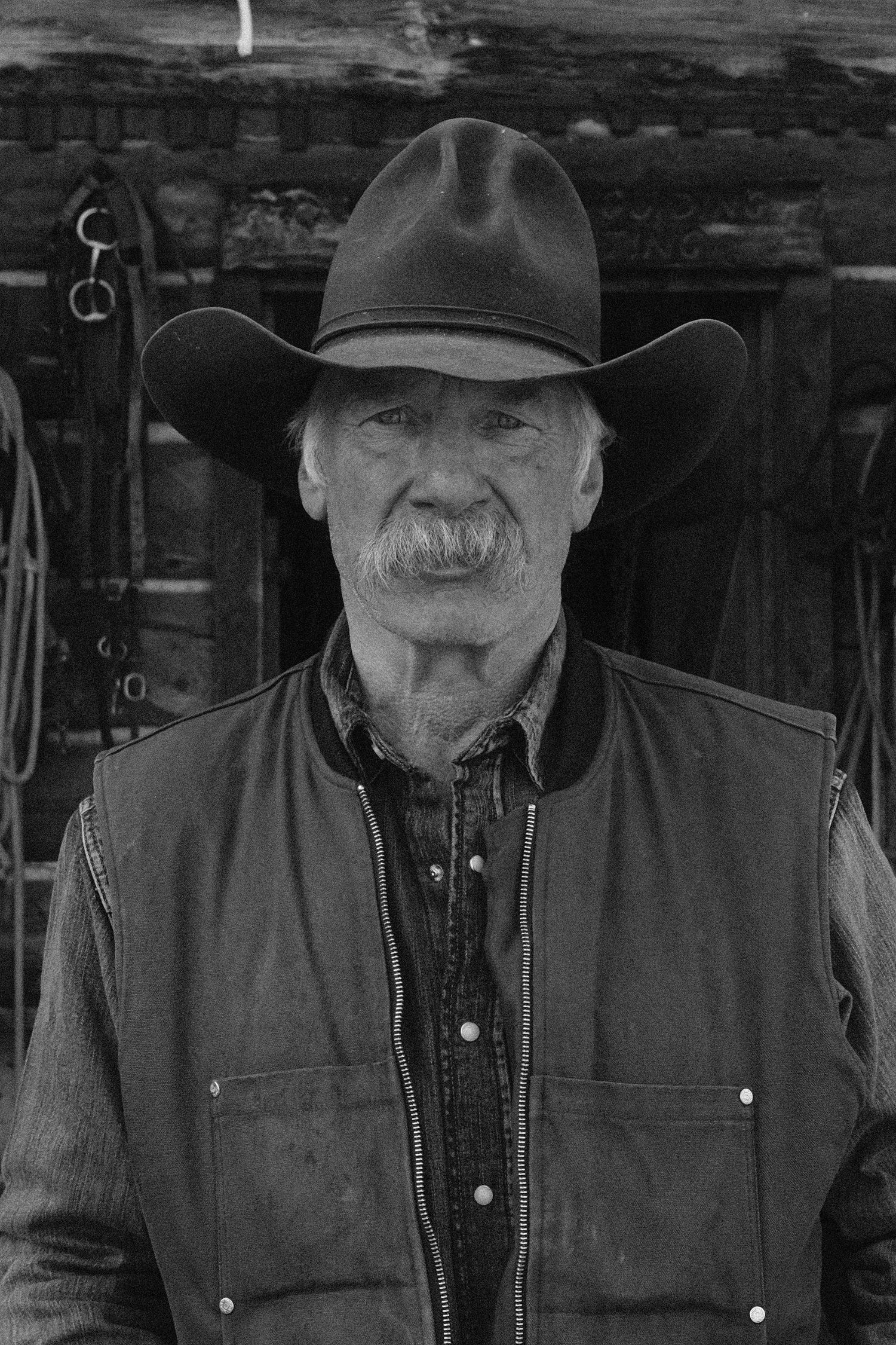 Black and white portrait of an elderly cowboy wearing a hat and vest, standing in front of a rustic barn.