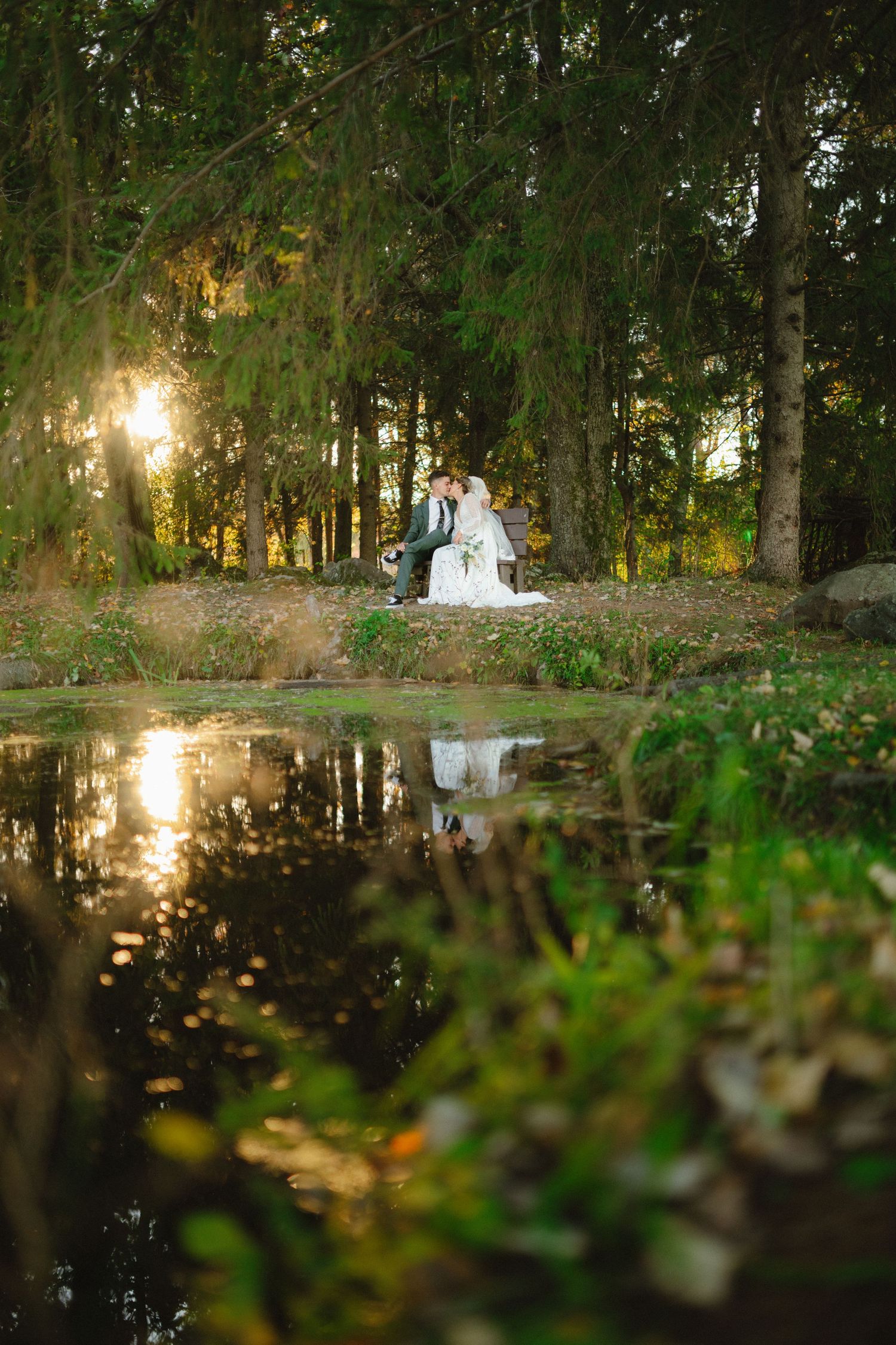 Wedding couple reflected in pond water surrounded by forest at sunset.