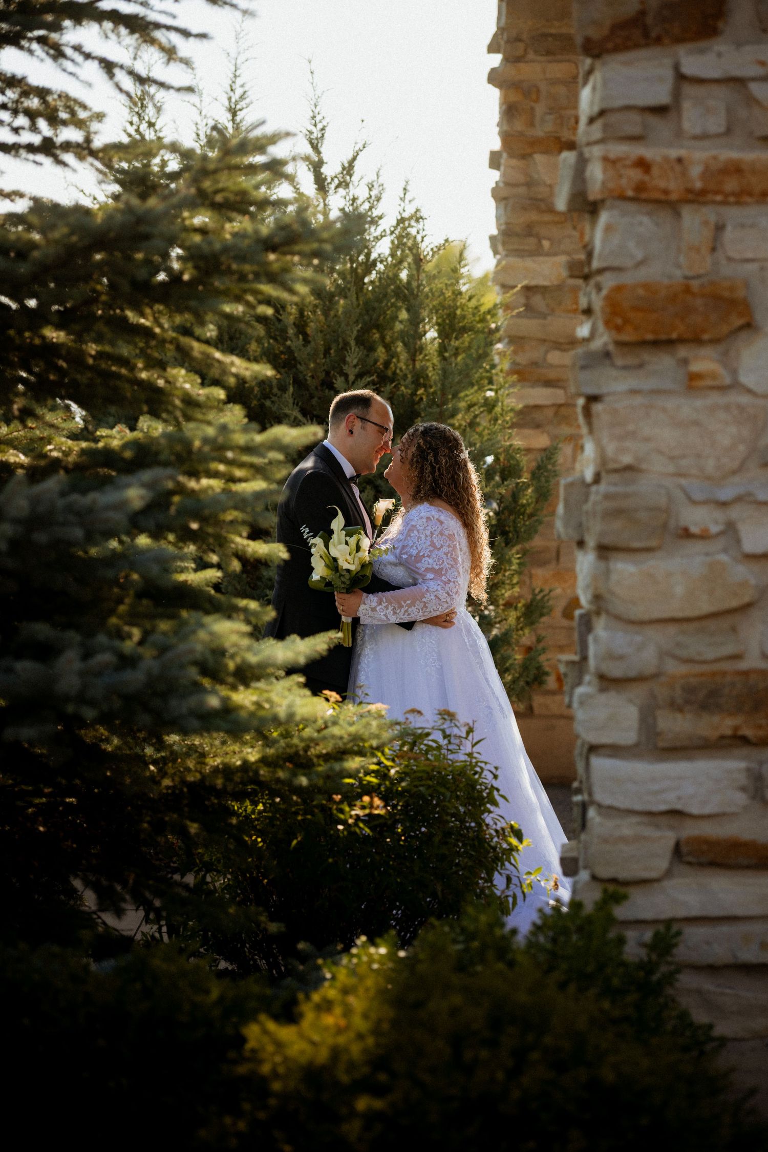 Couple embracing at sunset against rustic stone pillars and evergreen trees.