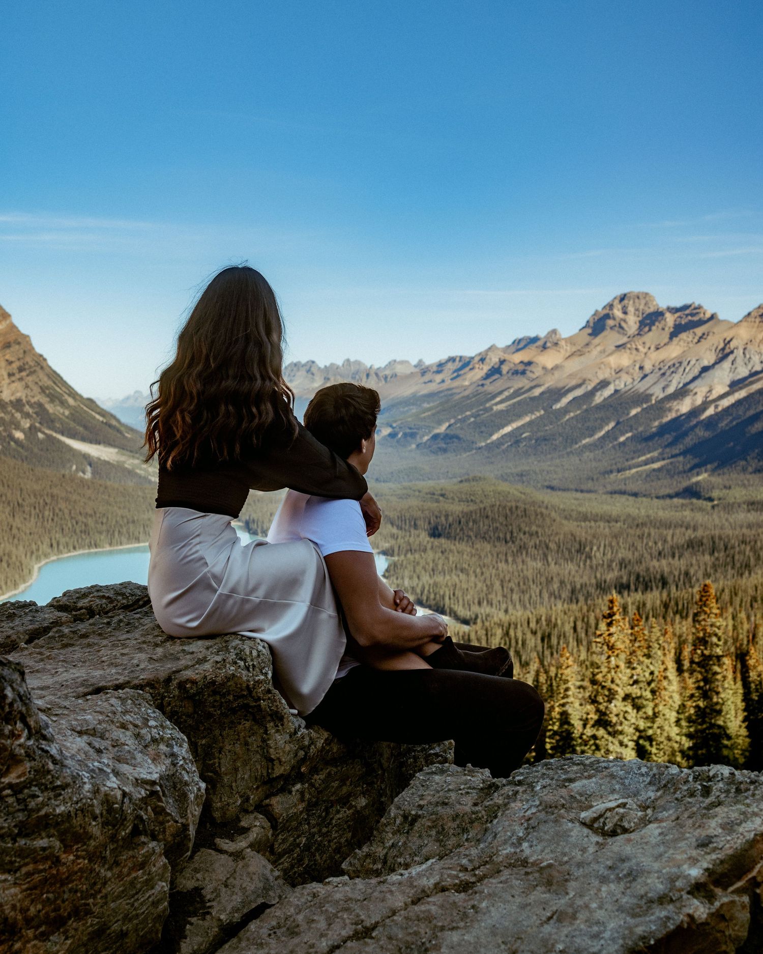 Two people sit on a rocky overlook enjoying panoramic mountain views with evergreen forests below.