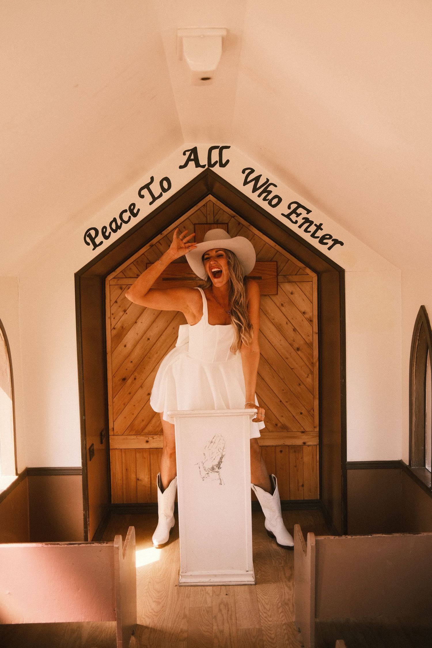 Woman in white dress, cowboy hat, and boots posing at chapel pulpit under Peace To All Who Enter sign.