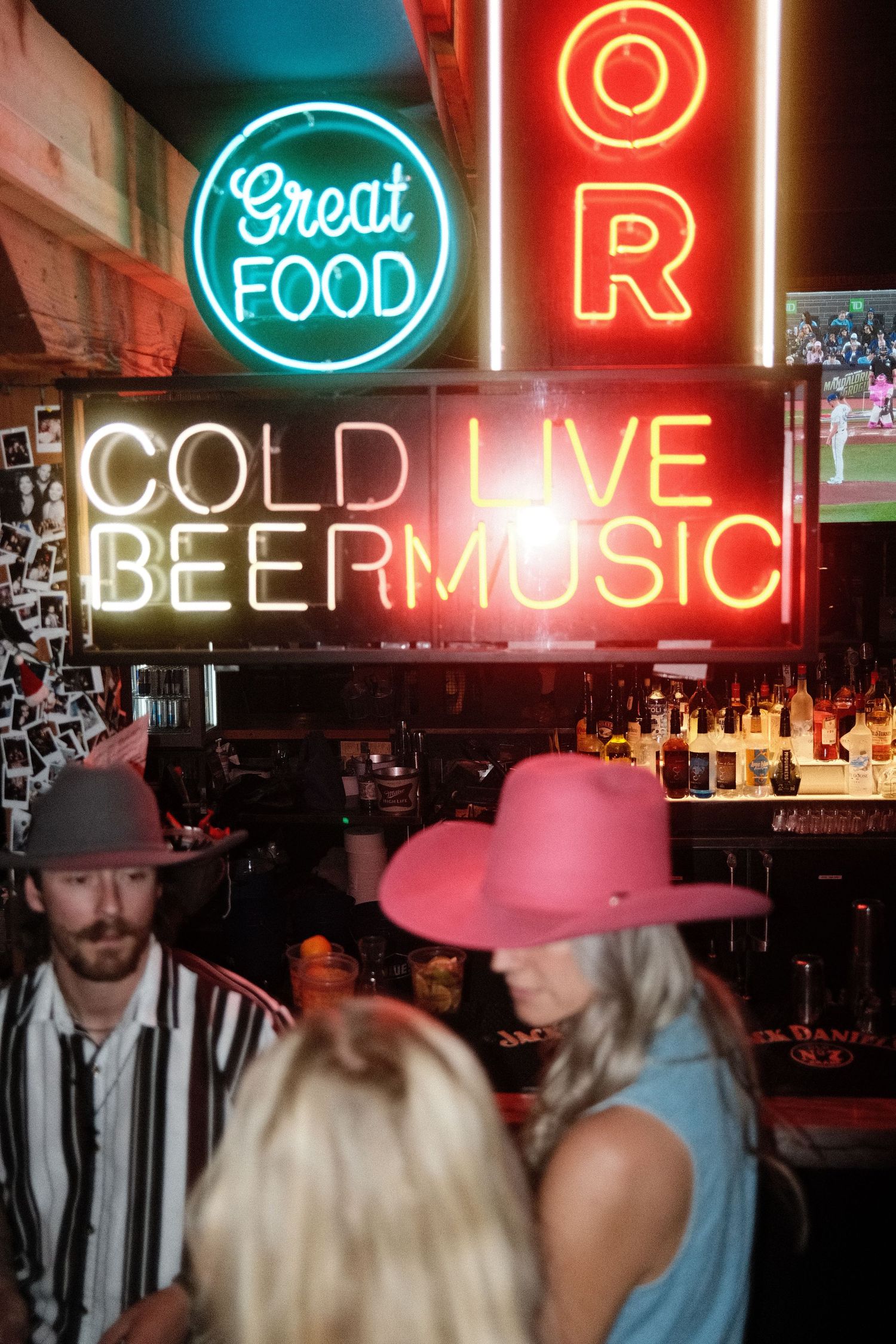 Neon signs reading Cold Beer, Live Music, and Great Food glow above patrons wearing cowboy hats at a lively bar.