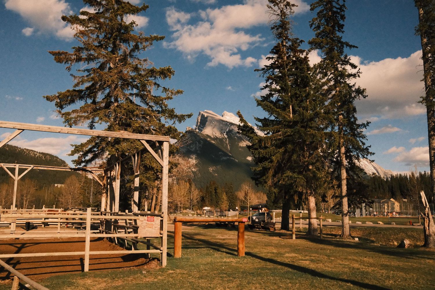 Rustic wooden structures and tall pine trees frame a stunning snow-capped mountain peak under a blue sky in Banff.