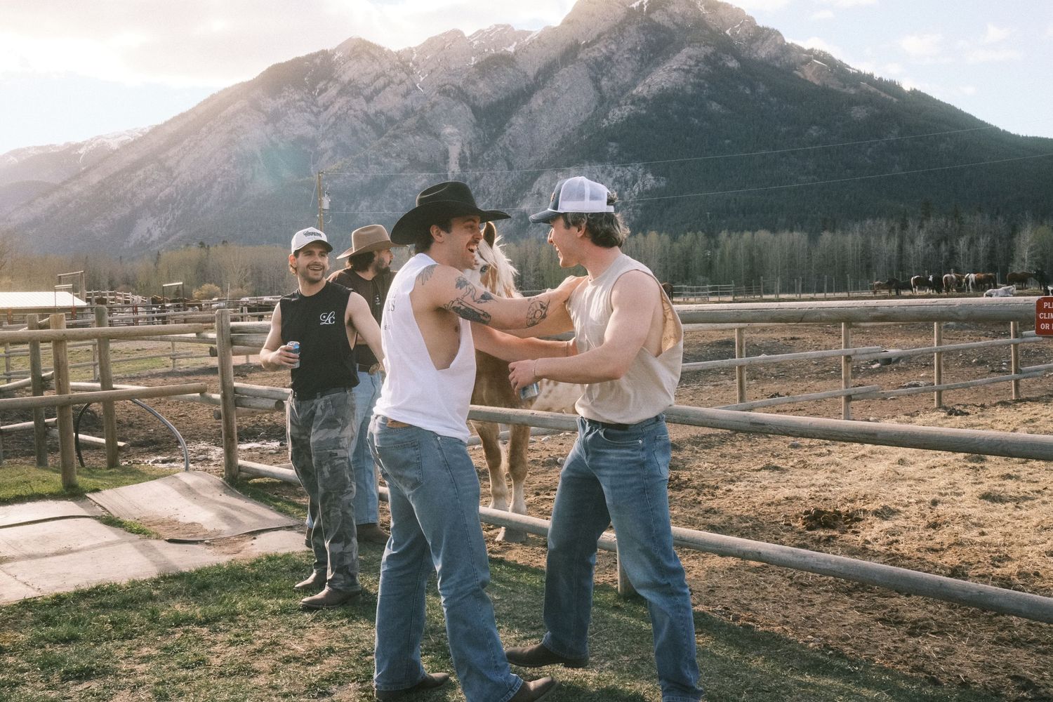 Two cowboys in jeans interact near a wooden fence with a mountain backdrop in a sunny outdoor ranch setting.