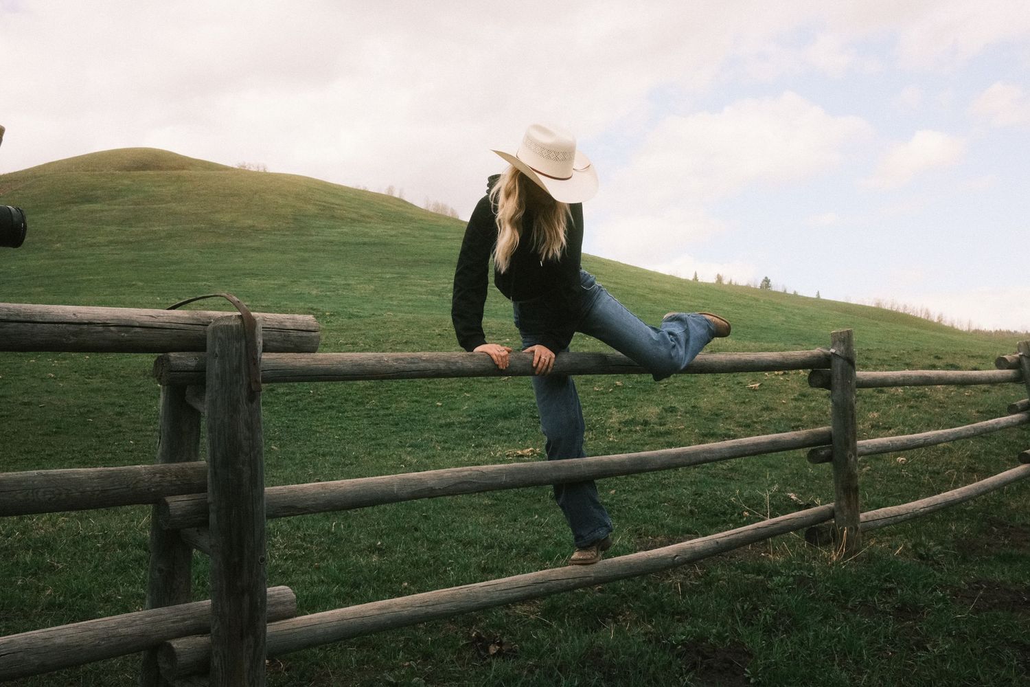 A woman in a white cowboy hat and jeans climbs a rustic wooden fence on a lush green hillside farm.