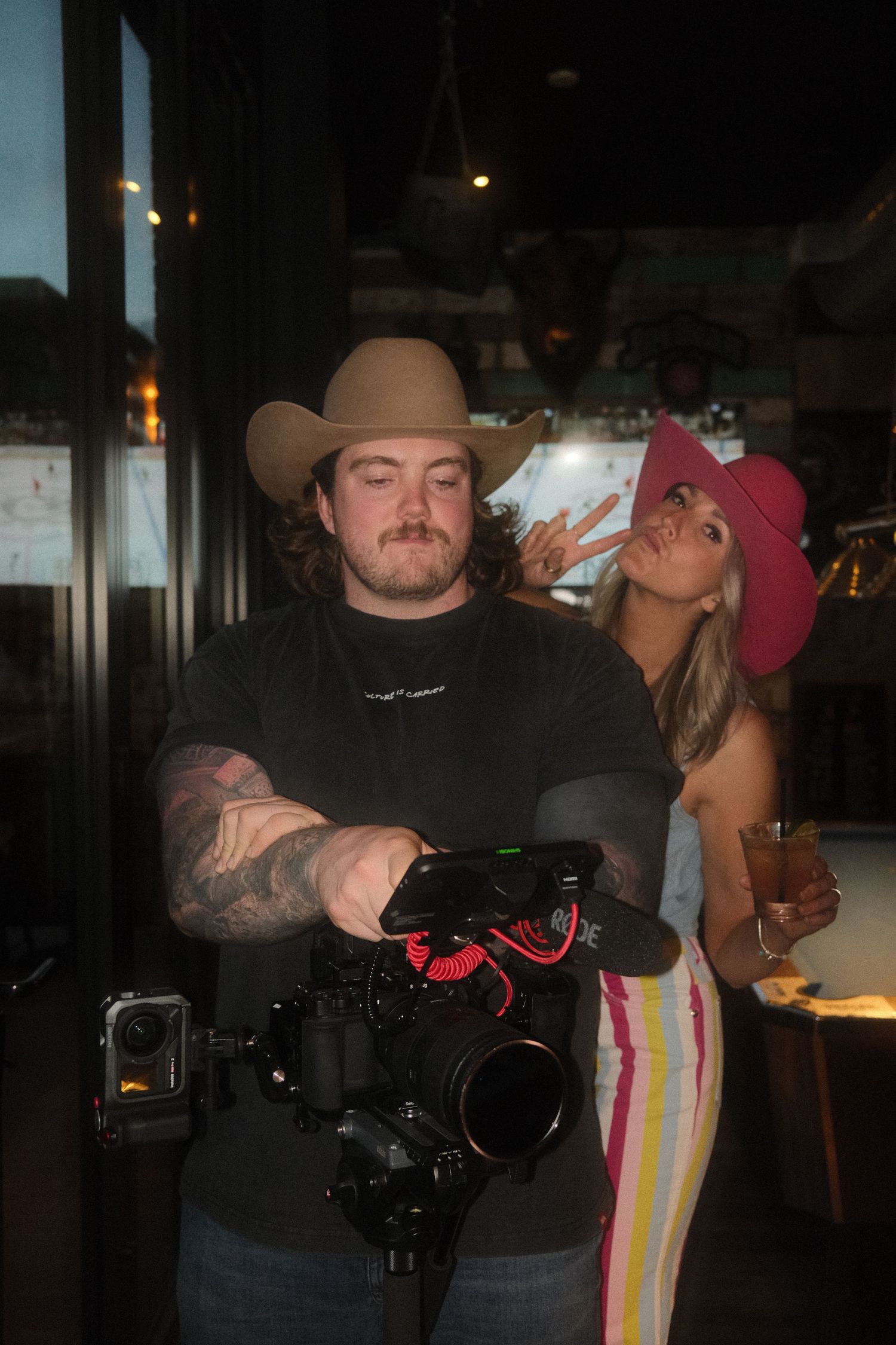 A cameraman in a tan cowboy hat holds a professional camera while a woman in a pink hat poses behind him indoors.