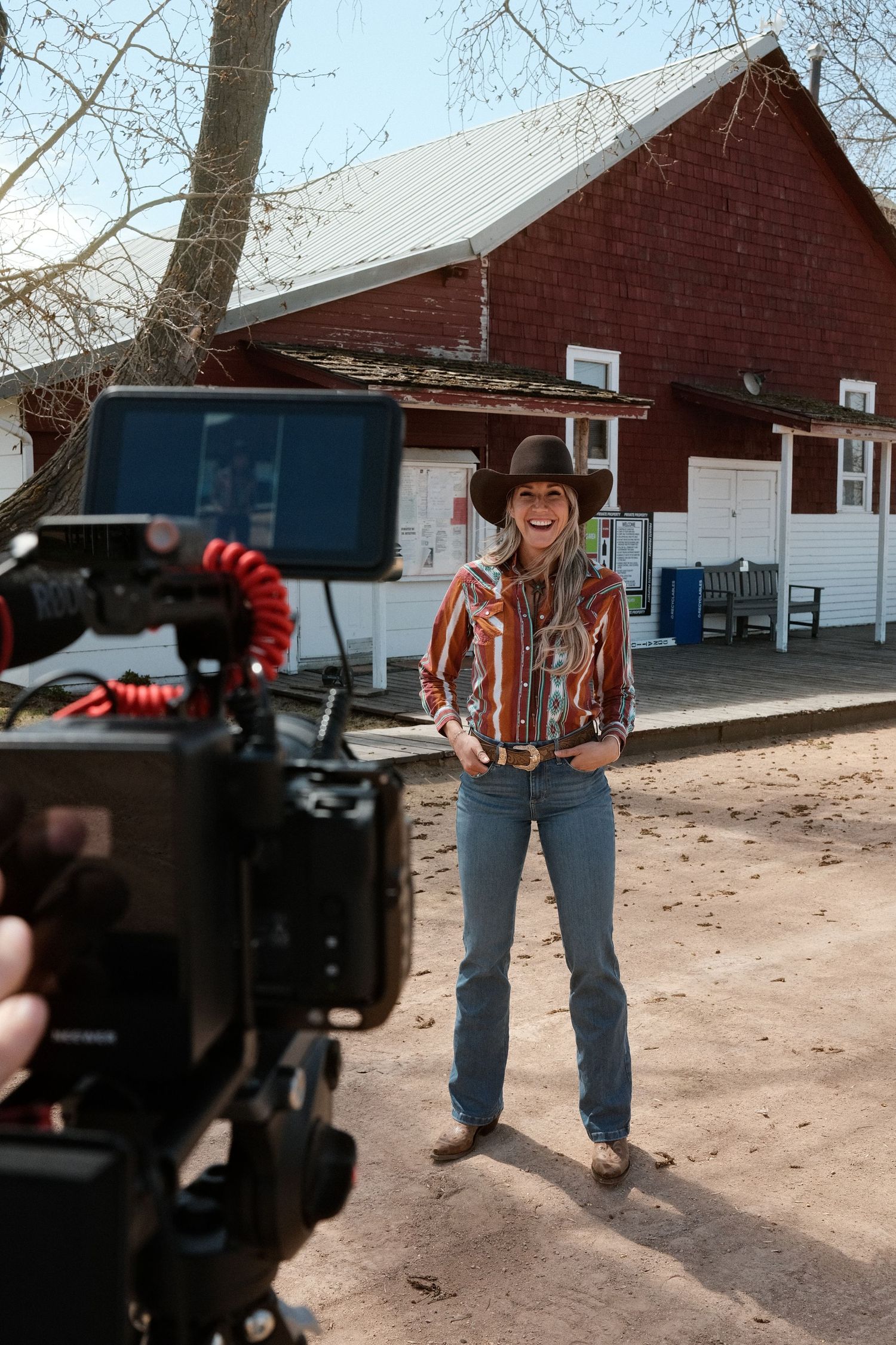 Woman in cowboy hat and plaid shirt smiling before a camera outdoors near a red barn.