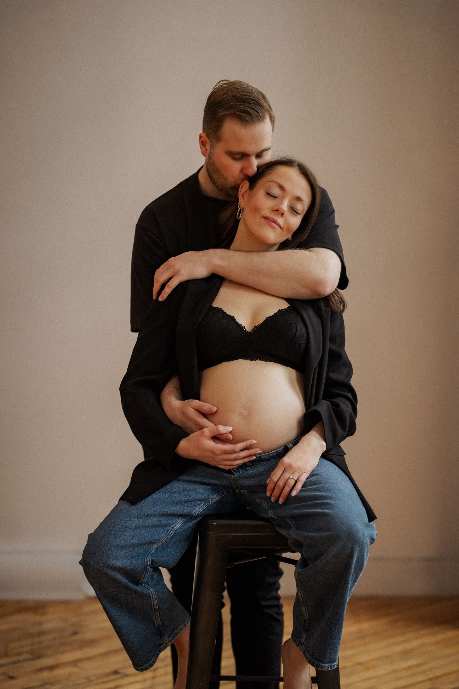 Intimate maternity session with couple sitting on wooden chair in natural light.