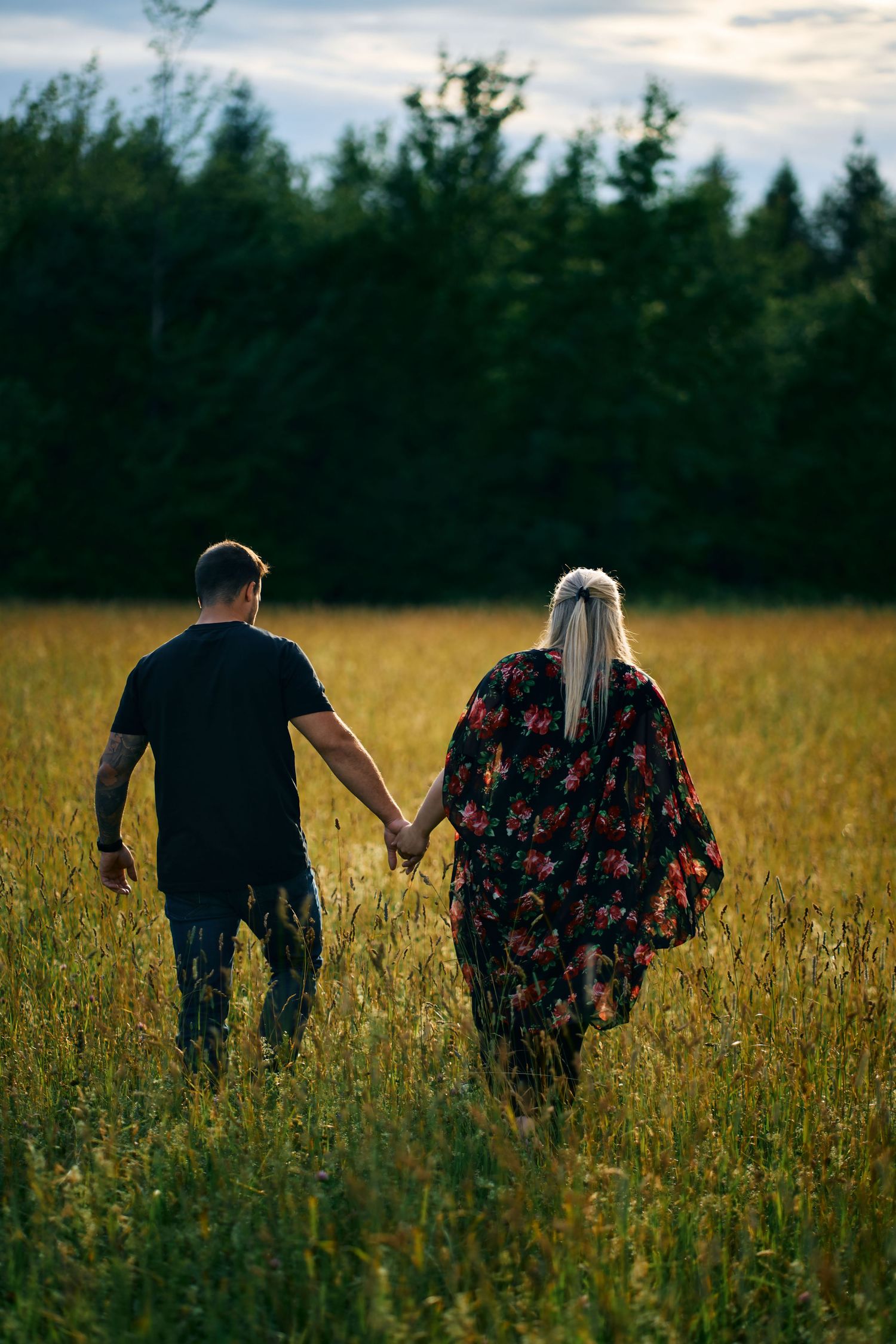 Couple walking hand in hand through a golden meadow at sunset with evergreen trees in background.