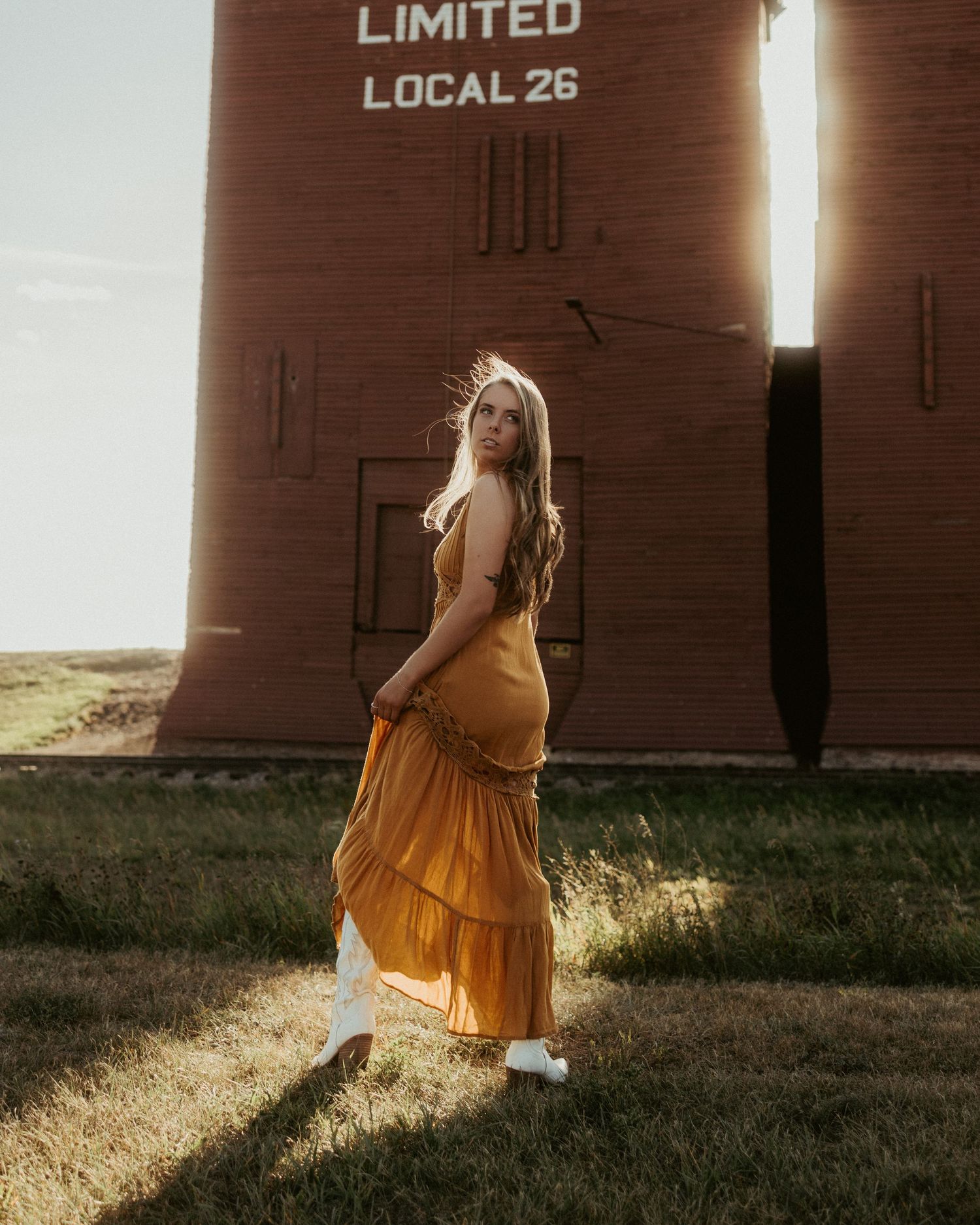 Figure in a flowing orange dress dances in front of rustic brown storage buildings at sunset.