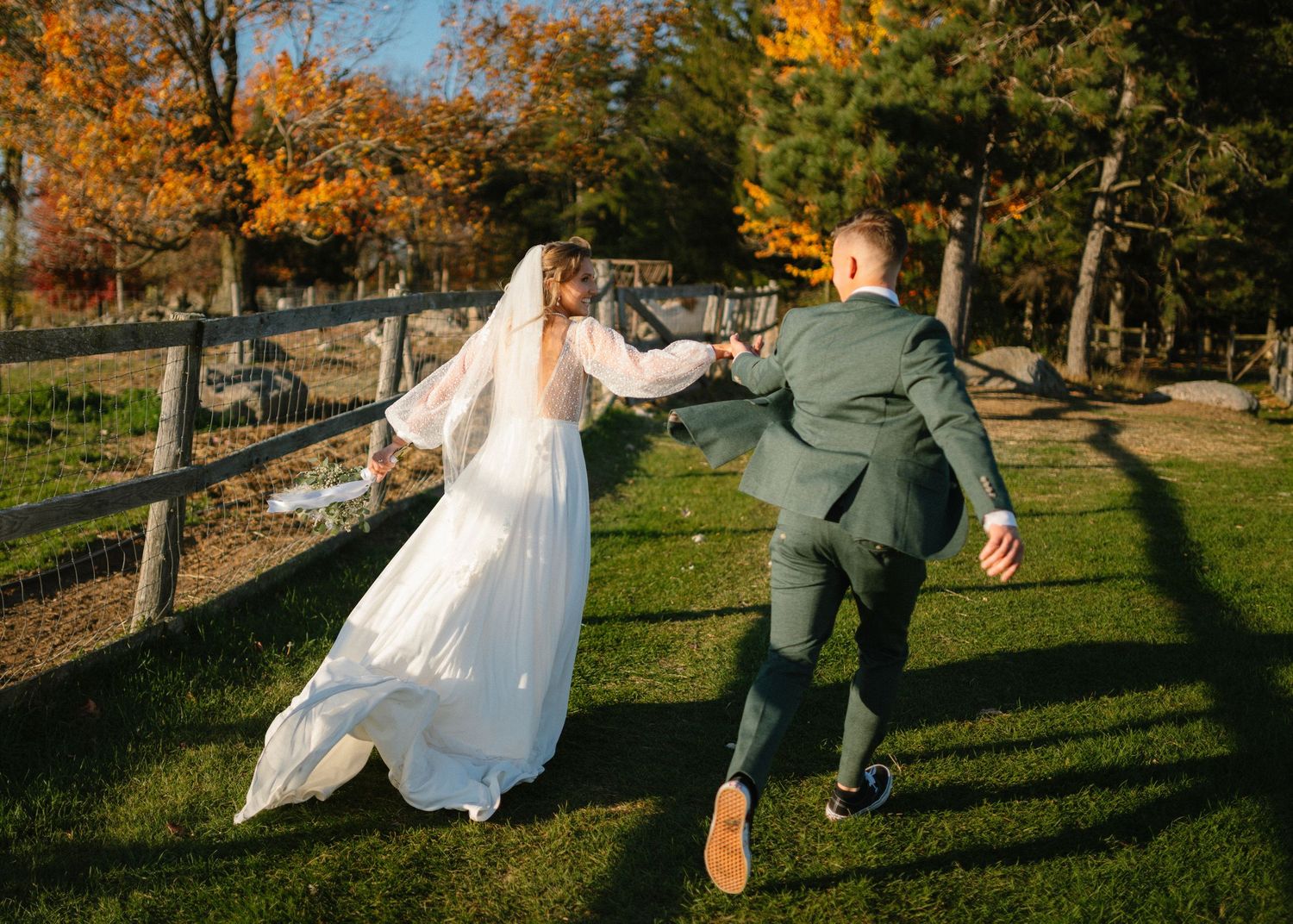 Playful wedding couple runs through autumn field with wooden fence.