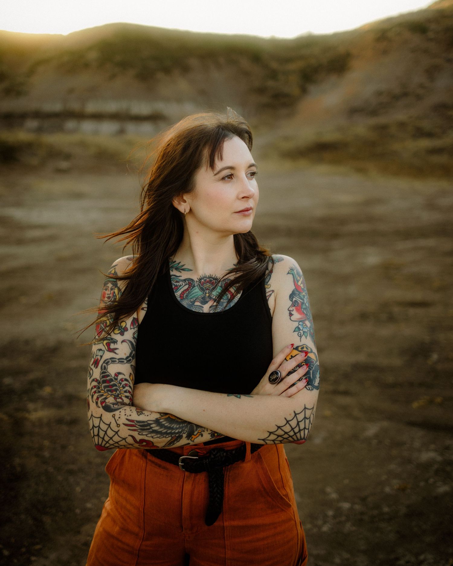 Woman in black top and rust-colored pants poses outdoors at sunset.