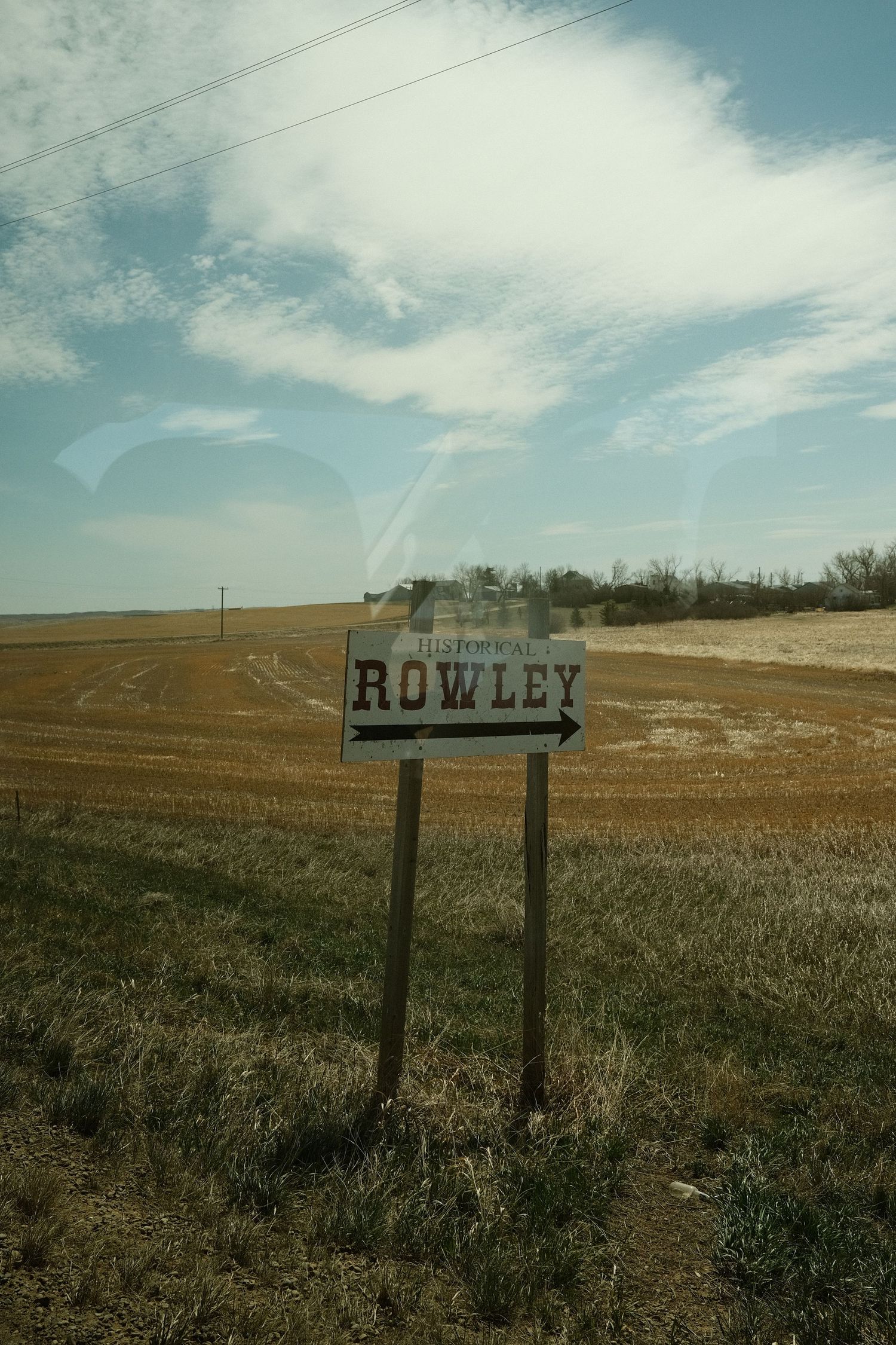 Rustic wooden sign pointing to Rowley along a rural highway surrounded by flat farmland under a partly cloudy sky.