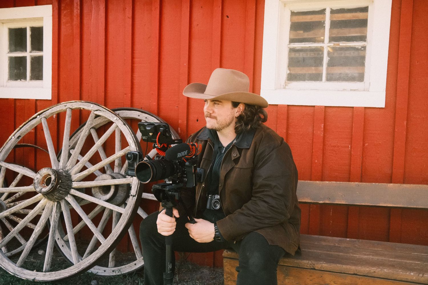 Filmmaker operating a camera near vintage wagon wheels beside a red wooden barn.