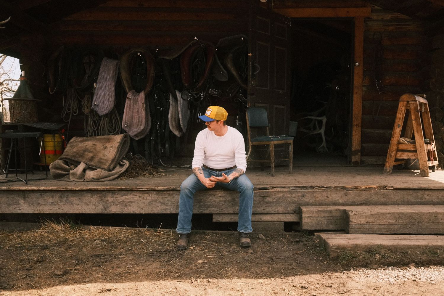 Man in white shirt and blue cap sits on rustic wooden porch steps near hanging leather tack and farm equipment.