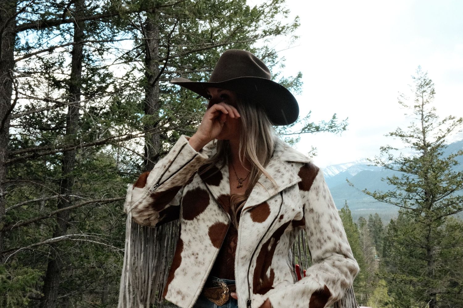 Woman in cowboy hat and cowhide fringe jacket poses outdoors among pine trees with mountain backdrop.