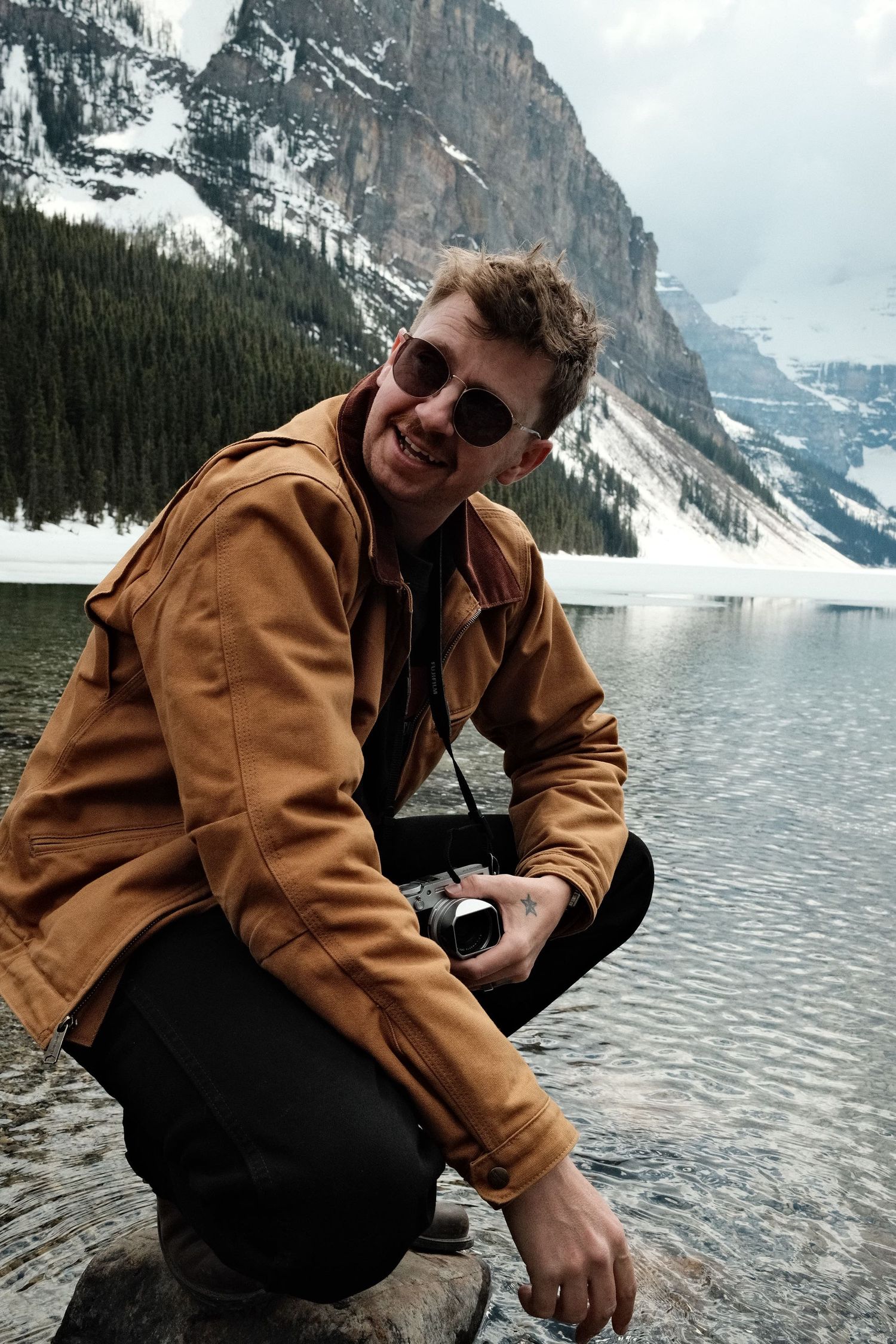 Smiling man in brown jacket and sunglasses crouching by a mountain lake with snow-capped peaks and pine trees.