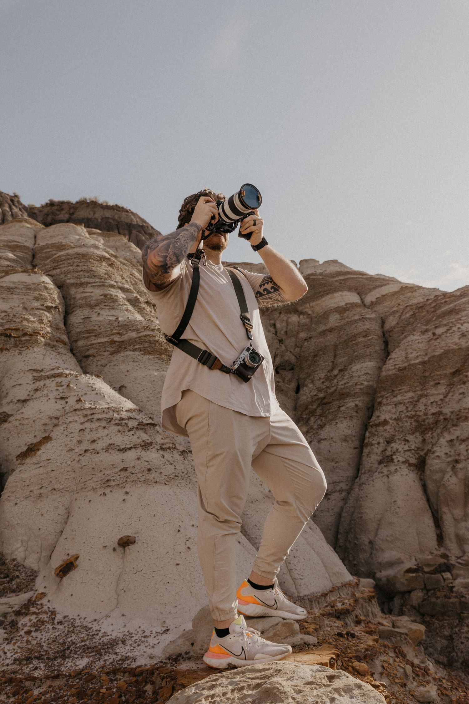 Tattooed male photographer shooting upward with telephoto lens while standing on rocks in a desert landscape.