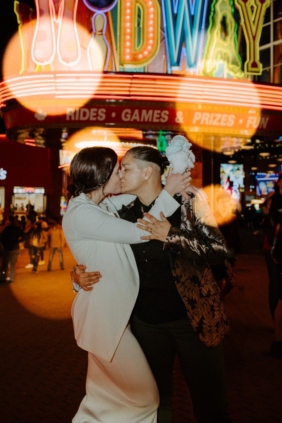 Couple embracing at night with colorful neon carnival lights in the background.