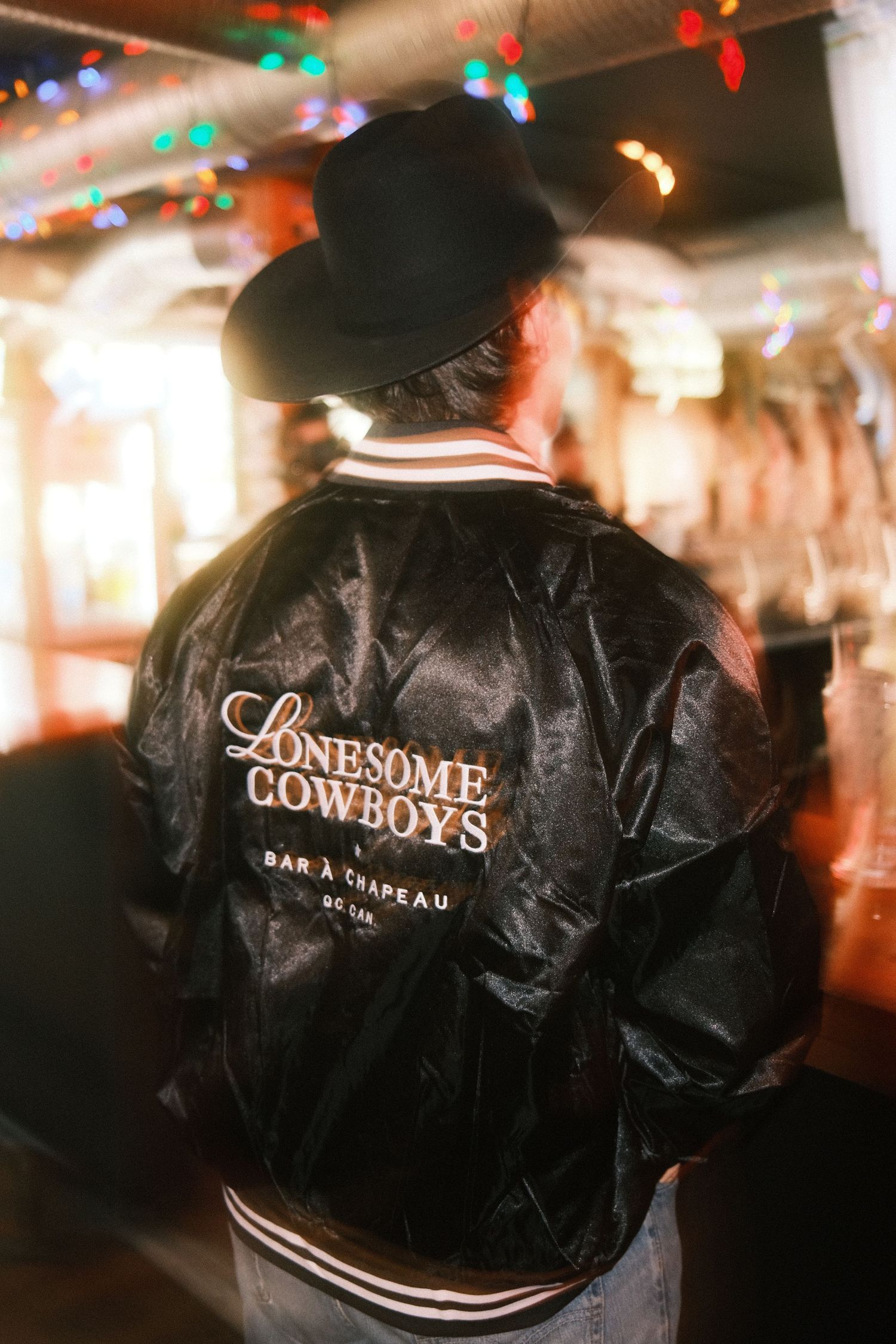 Person wearing a black Lonesome Cowboys Bar à Chapeau bomber jacket and cowboy hat in a festive bar setting.