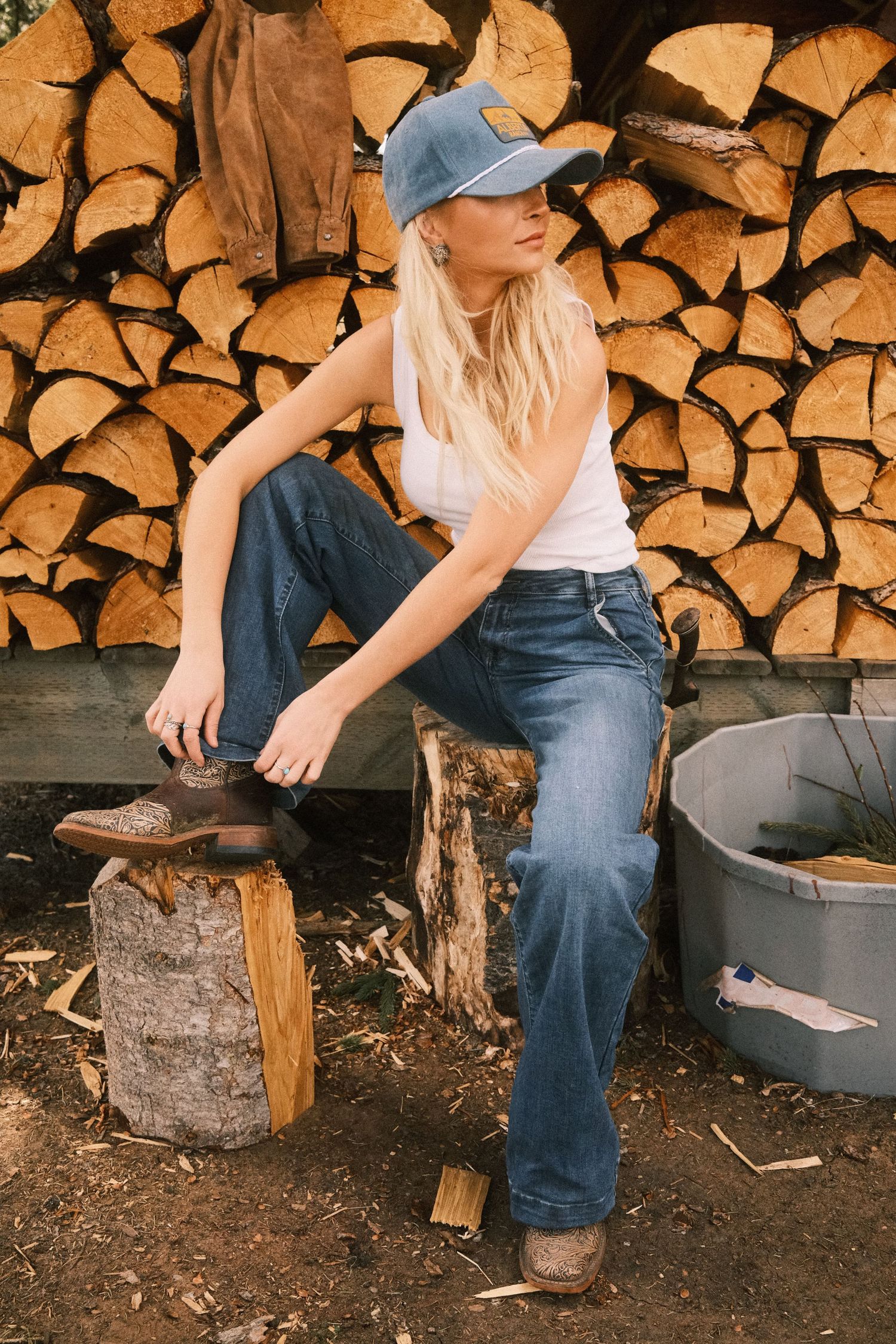 Blonde woman in white tank, blue jeans, and cap sits on log stump near stacked firewood pile outdoors.