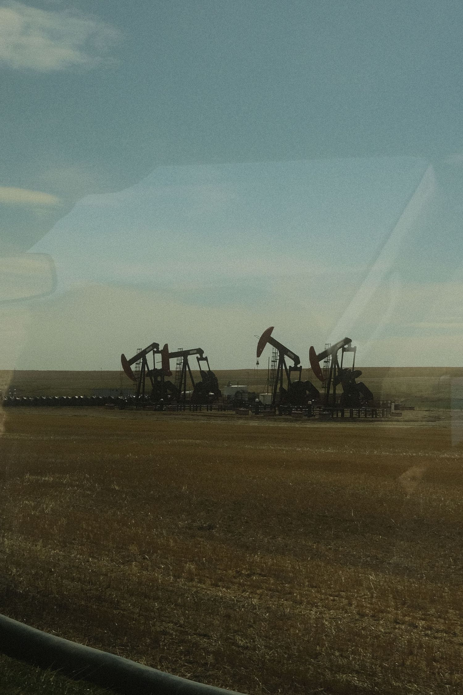 Multiple oil pump jacks operating in a flat prairie field under a partly cloudy sky, viewed through a vehicle window.
