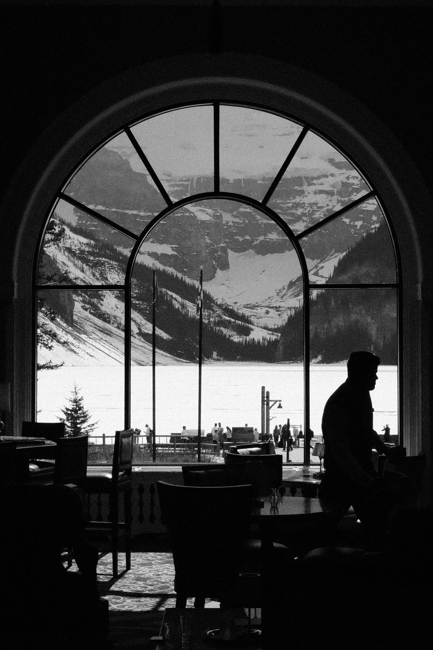 Black and white silhouette of a person standing by an arched window overlooking snowy mountains and a frozen lake.
