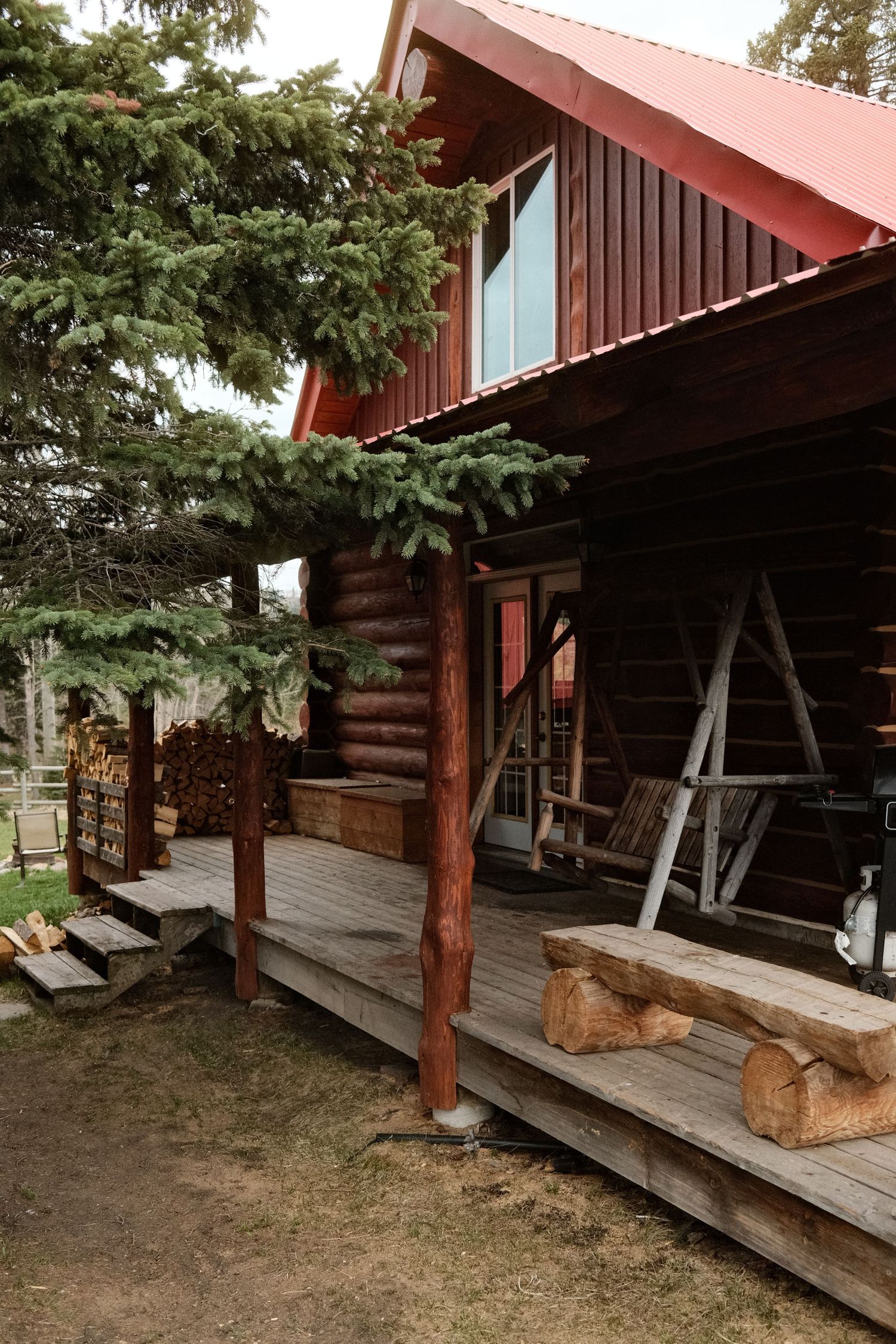 Rustic log cabin with red metal roof, wooden deck featuring log benches, and evergreen trees in a serene outdoor setting.
