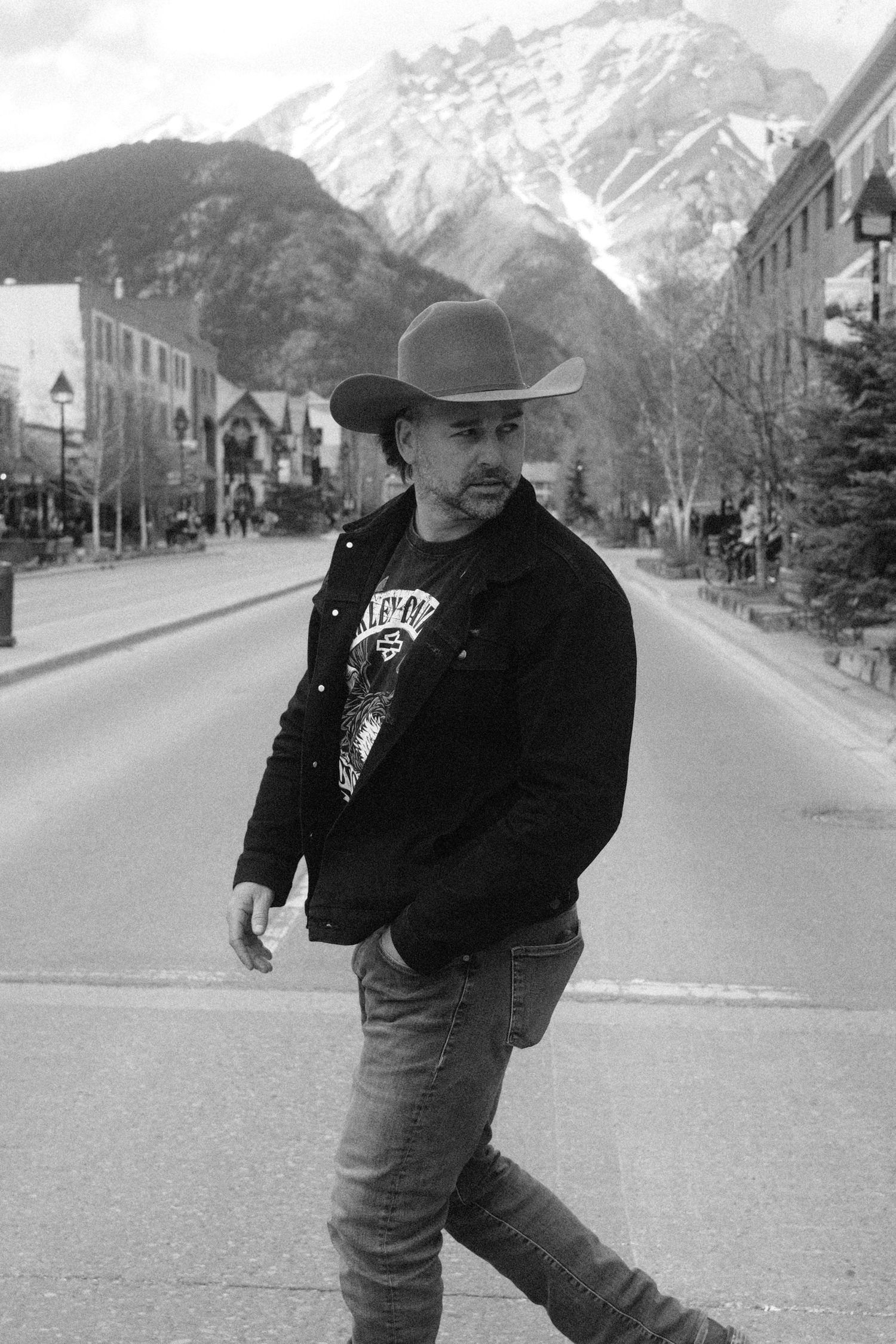 Black and white photo of a man in a cowboy hat and jacket walking on a mountain town street.