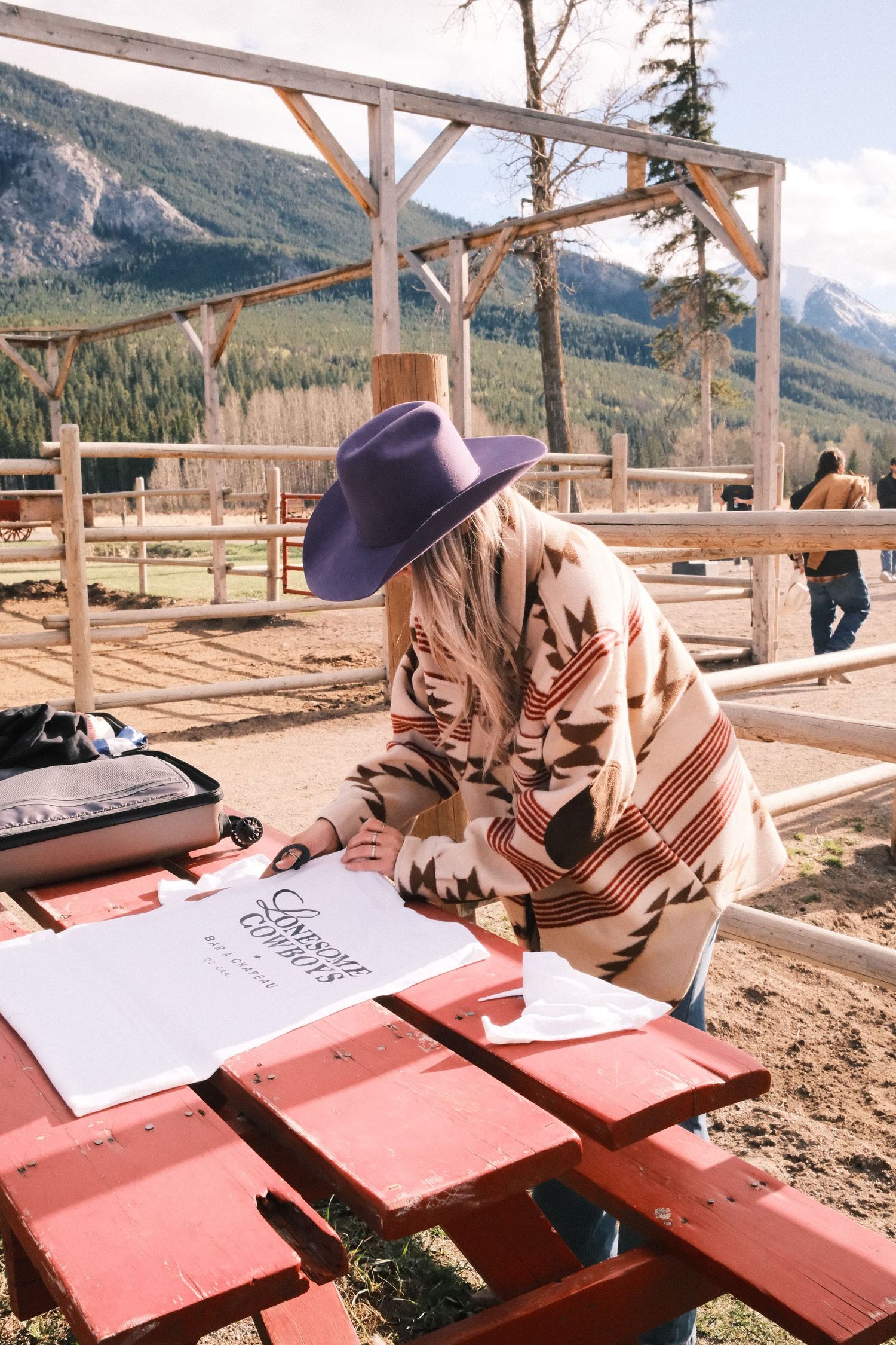 Woman in purple cowboy hat and Pendleton blanket coat reviews papers at a ranch with mountain views.