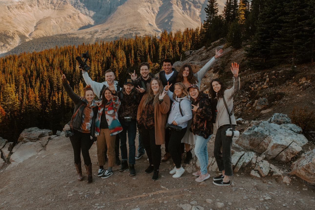 Group of friends posing and waving on a mountain trail surrounded by pine trees and rocky peaks during golden hour.