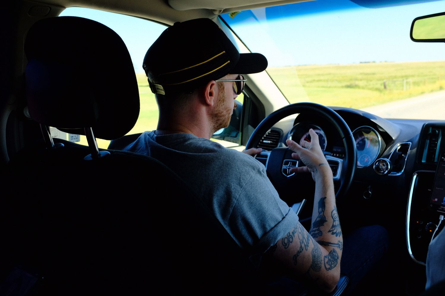 Tattooed man wearing a cap drives a car on a sunny highway, viewed from the backseat perspective.
