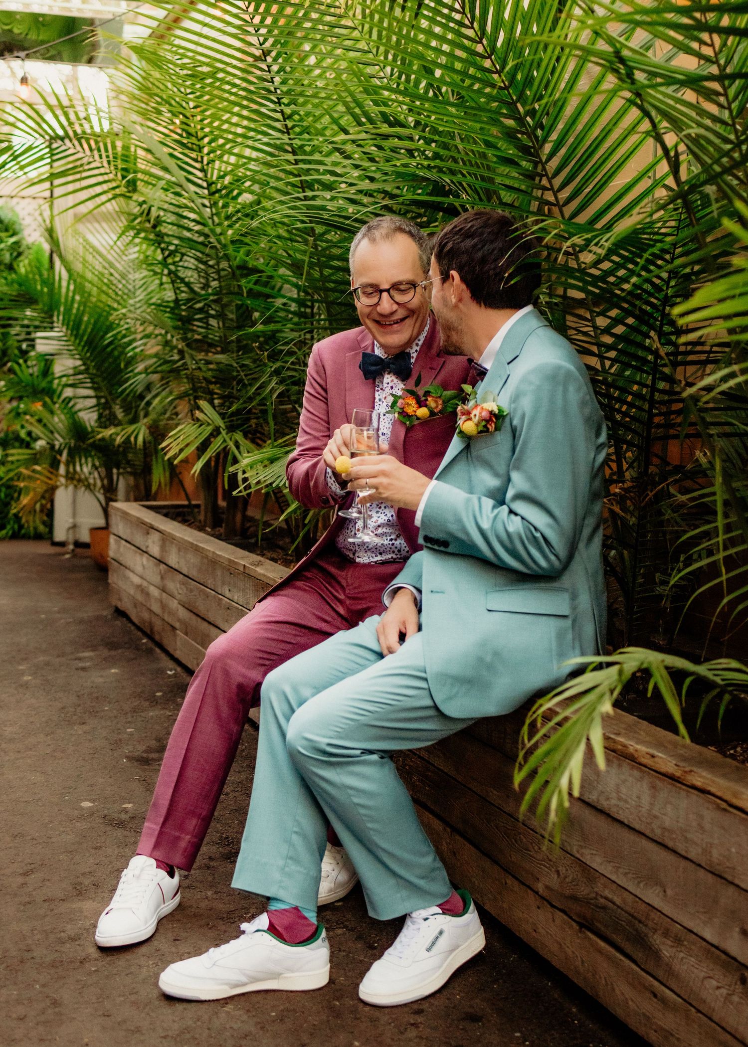 Couple in contrasting burgundy and mint suits sitting on wooden bench surrounded by palm fronds.
