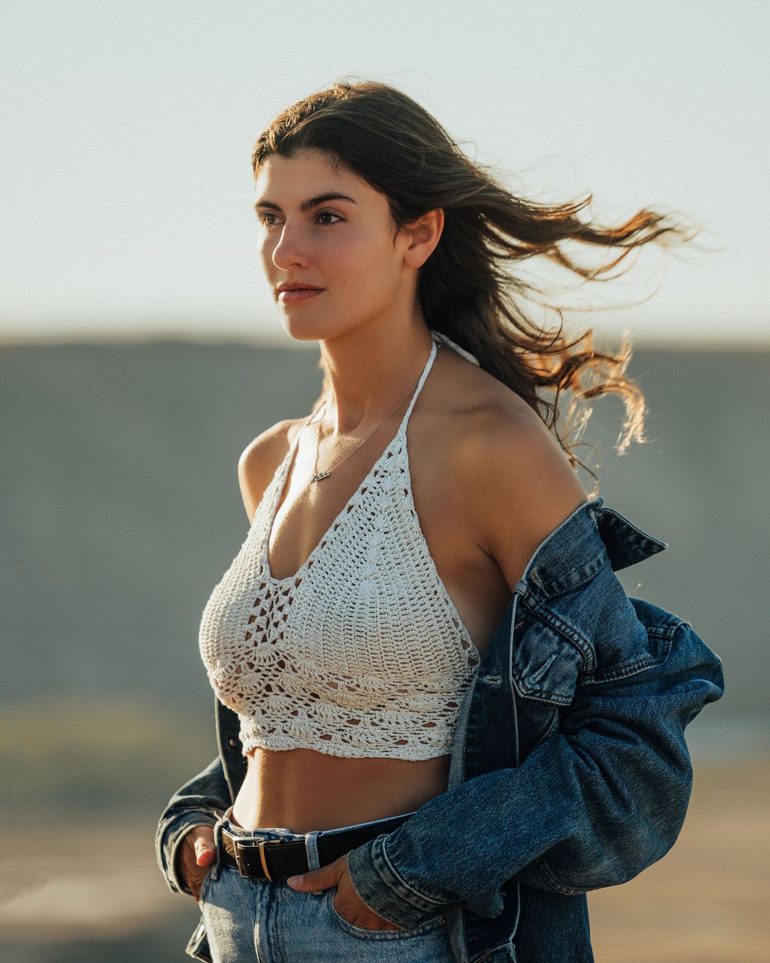 Model wearing white crochet top and denim jacket poses in desert landscape at sunset.