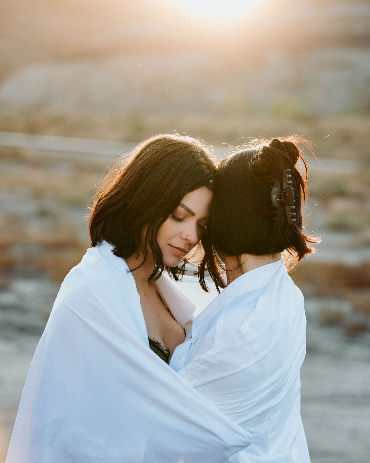 Couple draped in white fabric embraces against sunset backdrop.