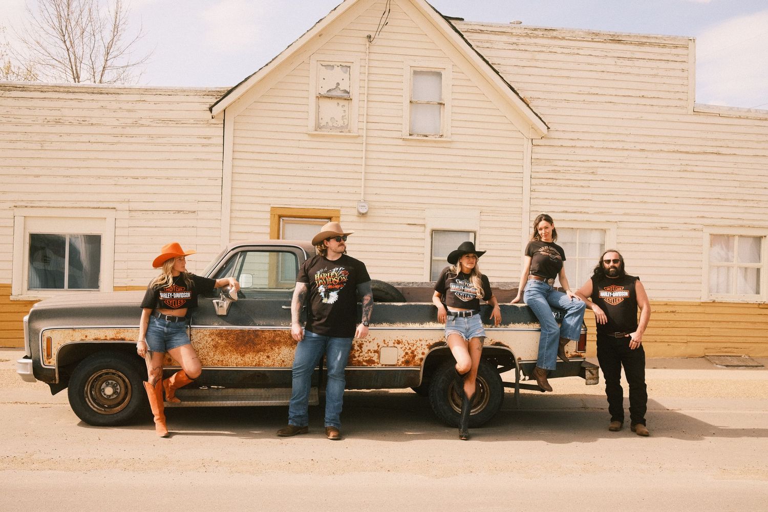 Country band posing with a rusty vintage pickup truck in front of a farmhouse on a sunny day.