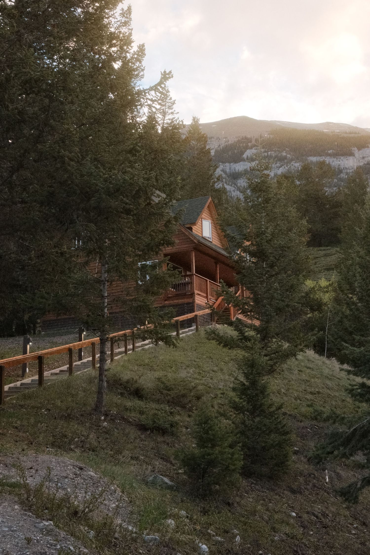 Rustic wooden cabin nestled among tall pine trees on a mountain hillside at sunset with rocky peaks in the background.