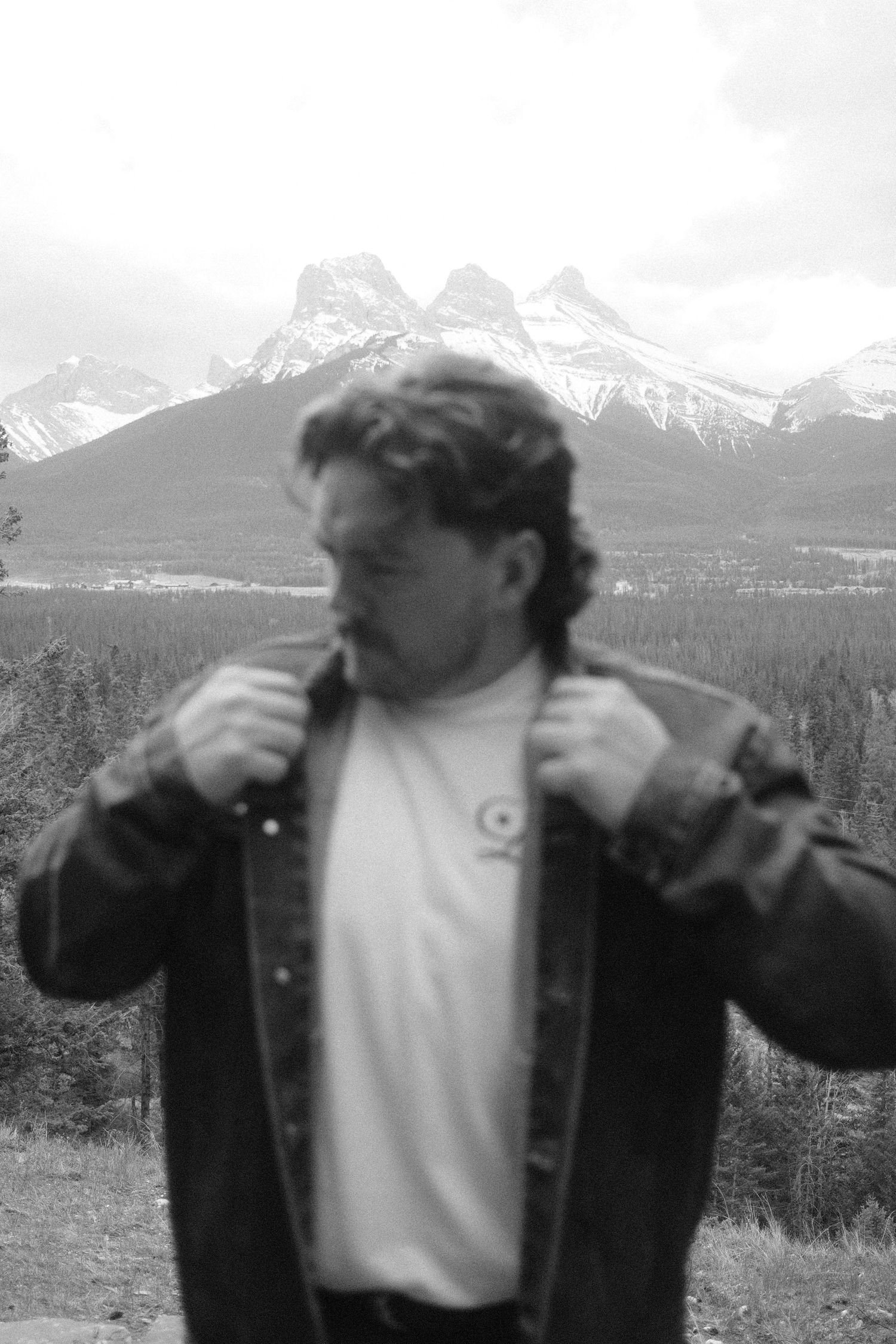 Black and white photo of a man adjusting his jacket collar with snow-capped mountains and forest in the background.